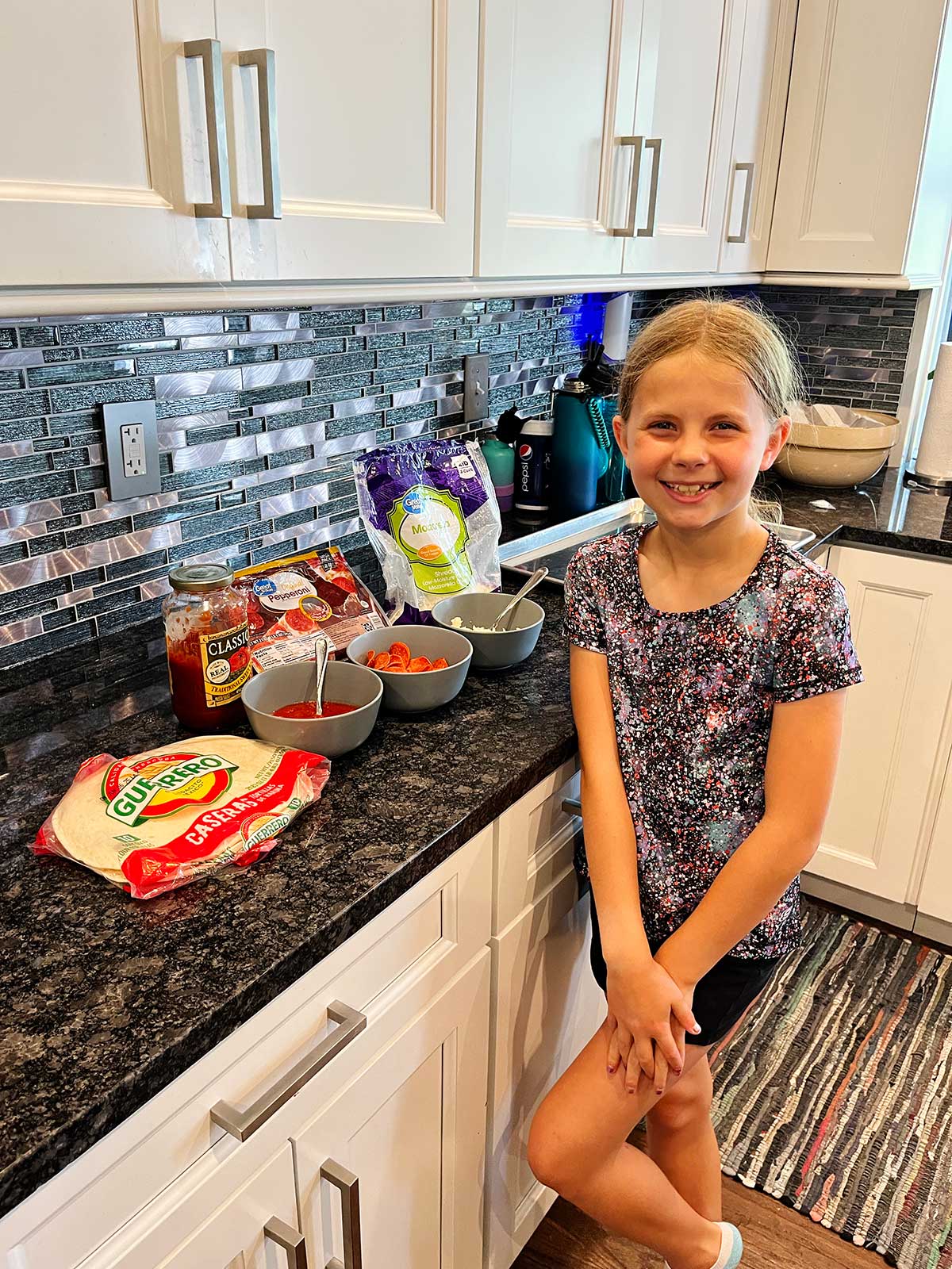 Uma menina jovem posando em frente a todos os suprimentos necessários para uma quesadilla de pizza na cozinha.