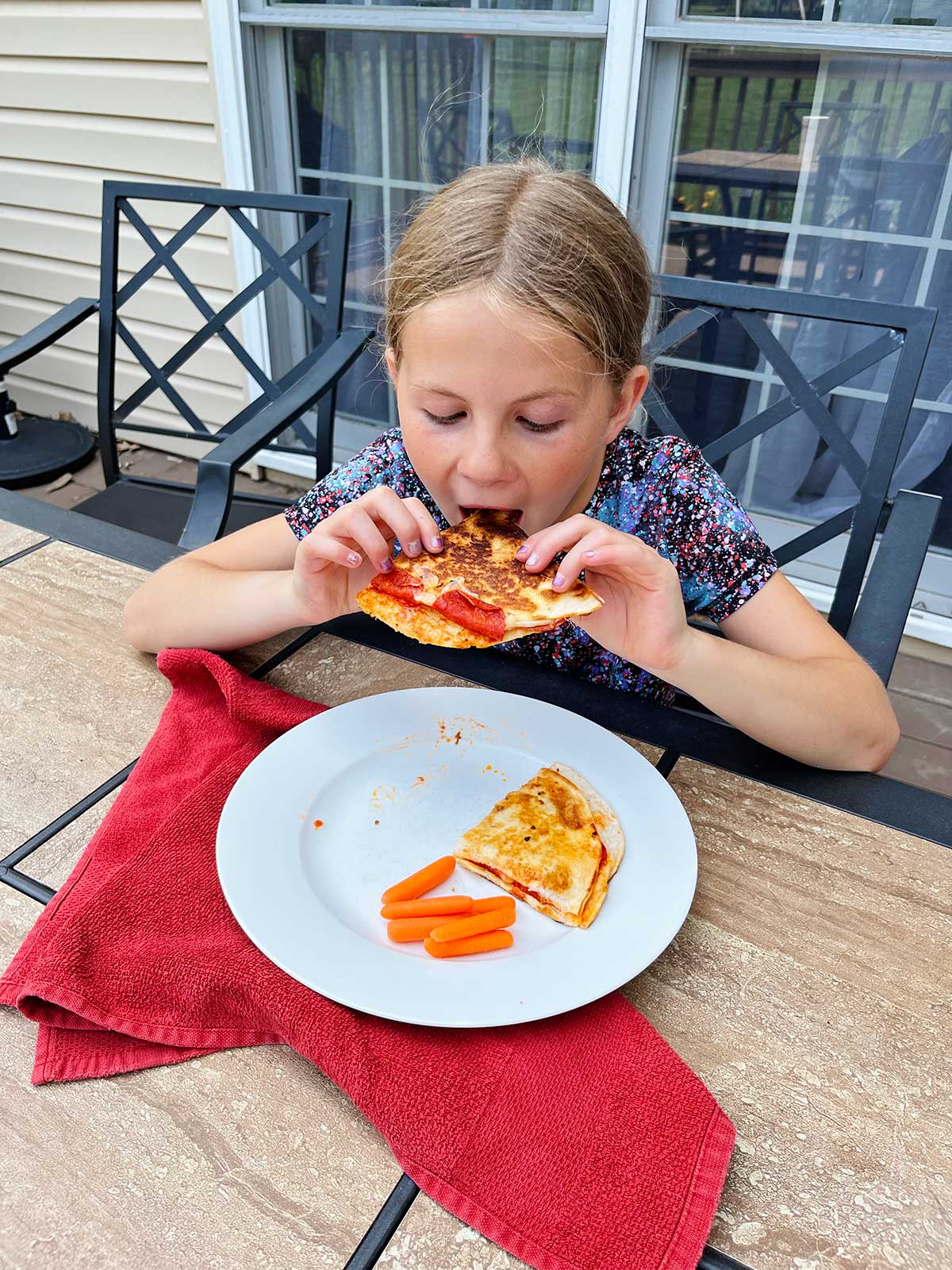 Uma menina jovem com cabelo loiro comendo uma quesadilla de pizza em uma mesa ao ar livre.