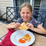 Uma menina jovem com cabelo loiro sorrindo para a câmera e segurando uma quesadilla de pizza em uma mesa ao ar livre.