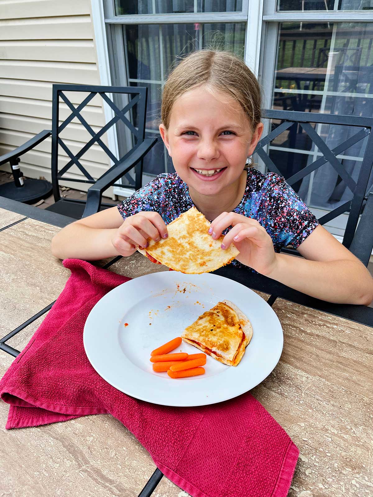 Uma menina com cabelo loiro sorrindo para a câmera e segurando uma quesadilla de pizza em uma mesa ao ar livre.