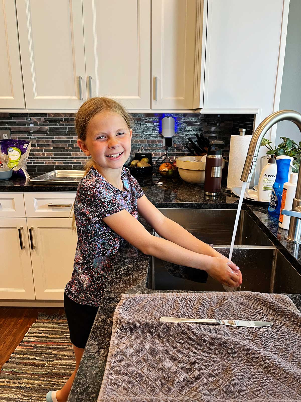 Uma menina com cabelo loiro lavando as mãos na pia da cozinha.