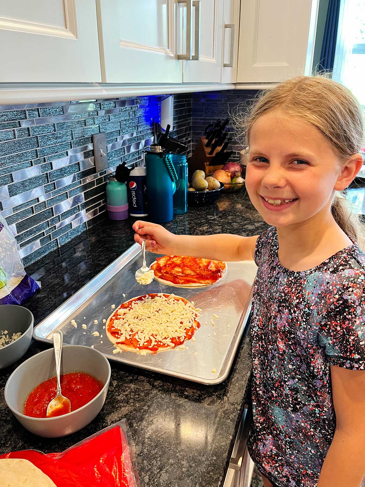 Uma menina jovem, sorrindo para a câmera, montando duas quesadillas de pizza na bancada.