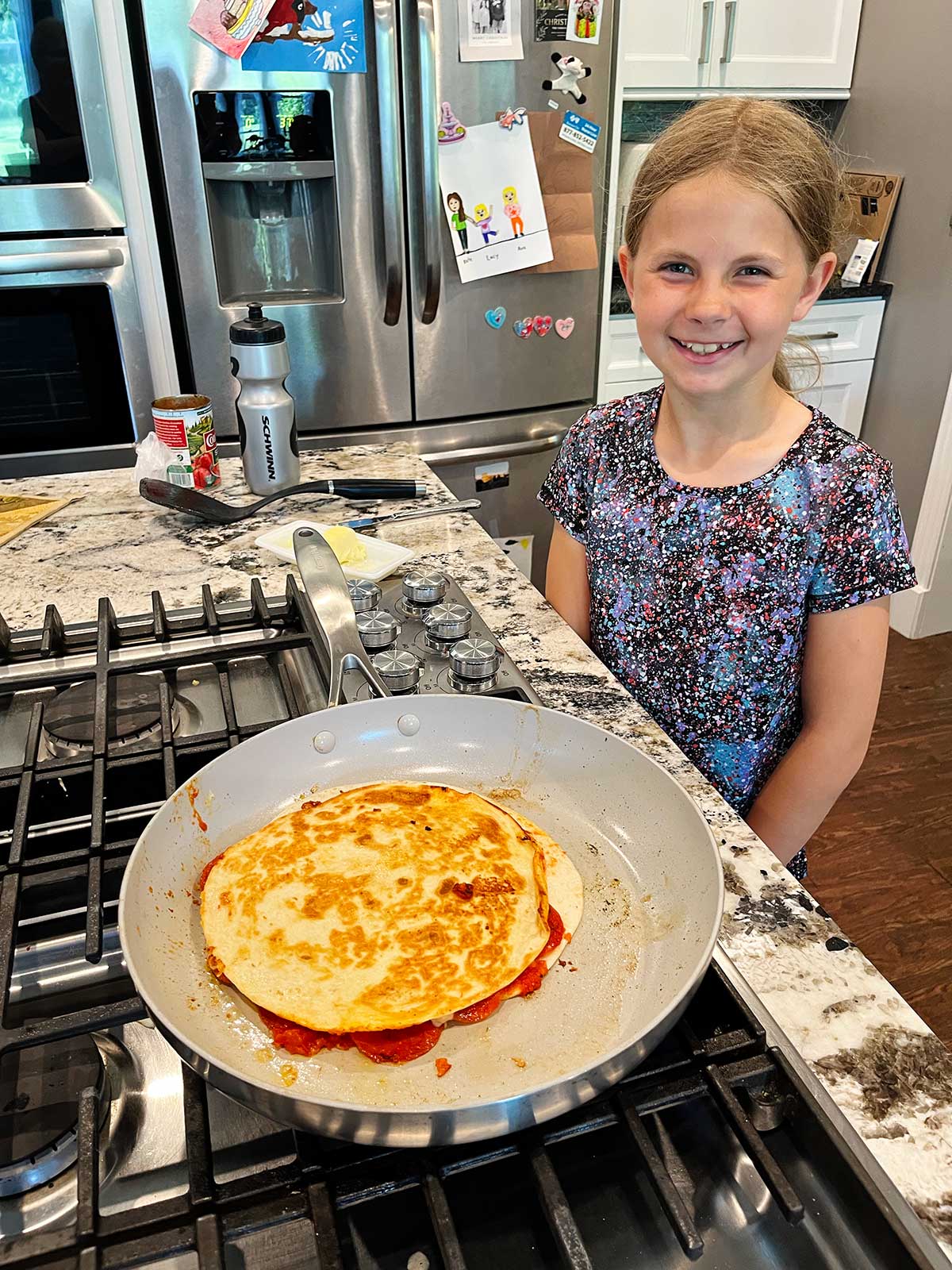 Uma menina jovem com cabelo loiro sorri para a câmera enquanto cozinha uma quesadilla de pizza no fogão.