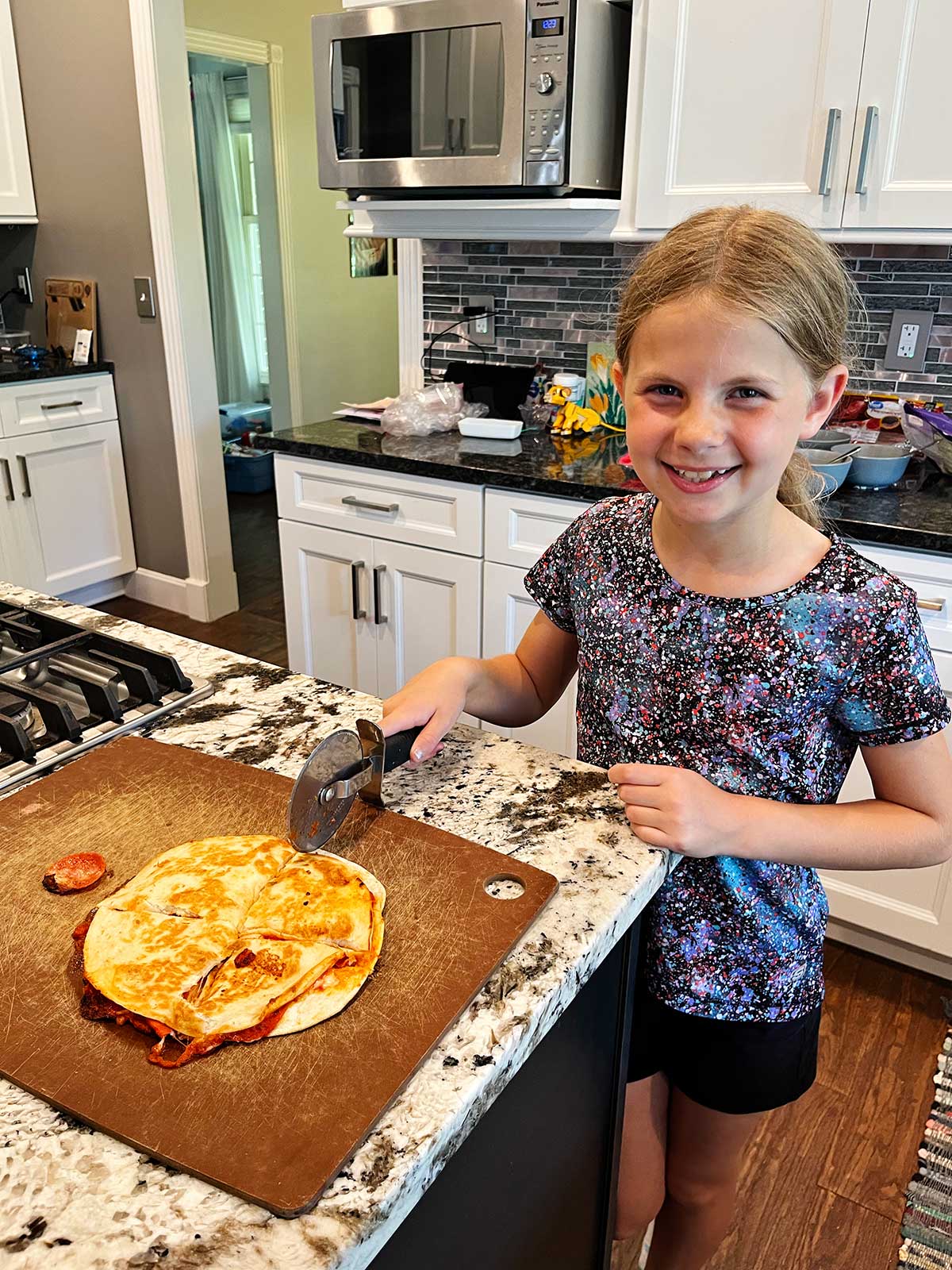 Uma menina jovem com cabelo loiro sorrindo para a câmera e cortando uma quesadilla de pizza em quatro partes na cozinha.