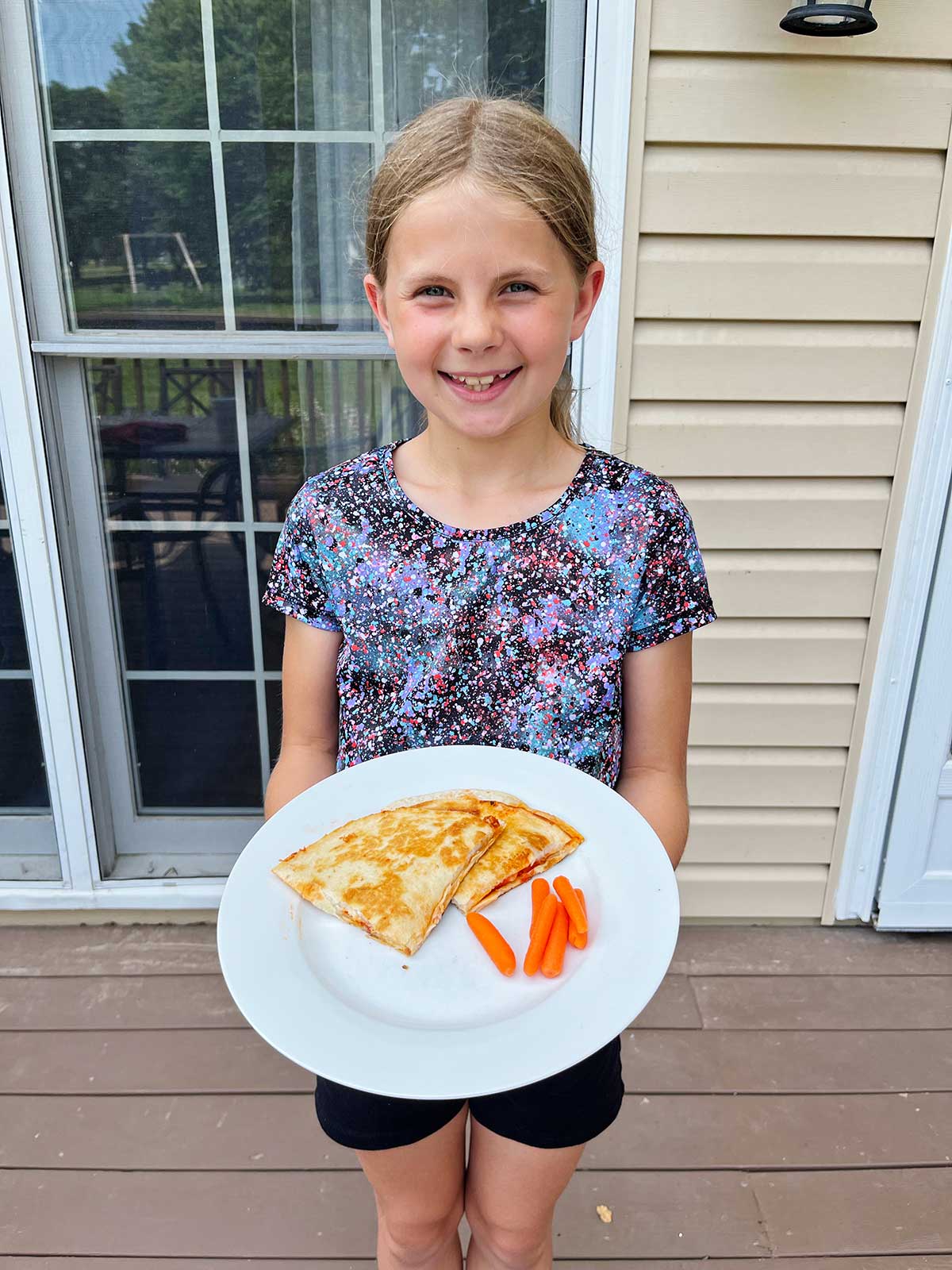 Uma jovem menina sorridente com cabelo loiro segurando um prato branco com duas quesadillas de pizza e cenouras baby do lado de fora.