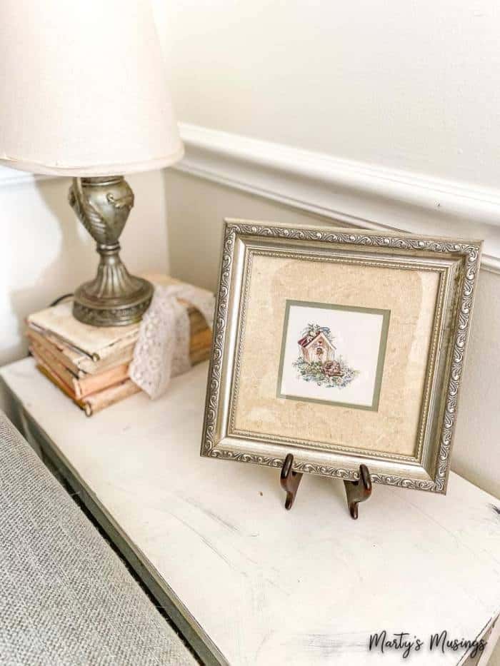 quadro emoldurado de uma casa sobre mesa lateral com livros pintados Um espelho sobre uma mesa