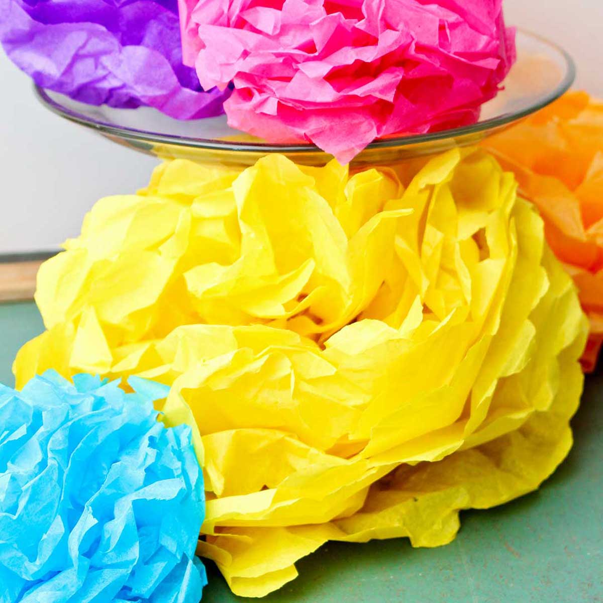 Flores de papel de seda coloridas concluídas, uma roxa e outra rosa descansando em um suporte de bolo de vidro com outras embaixo.