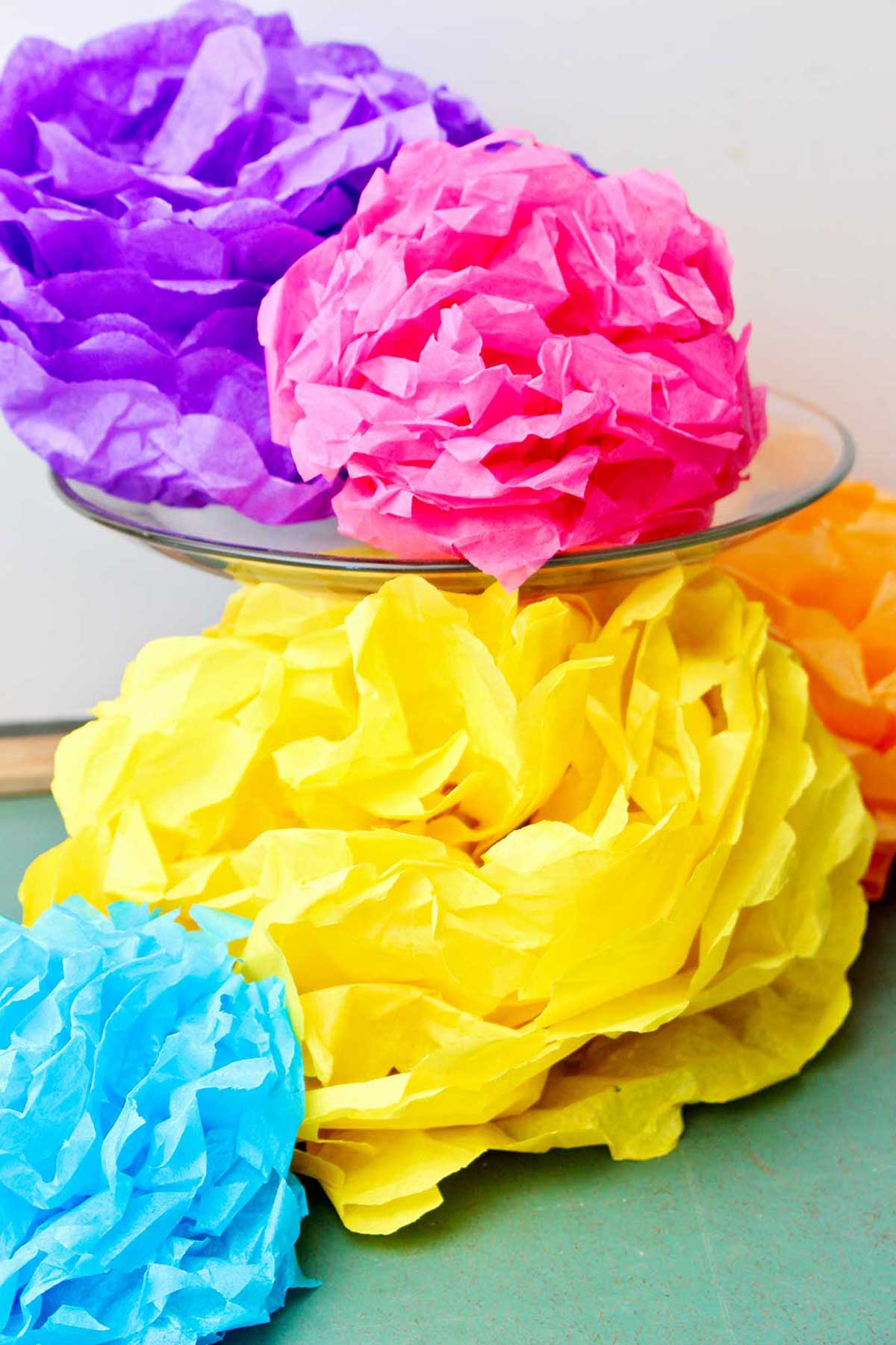 Flores de papel de seda coloridas concluídas, uma roxa e rosa descansando em um suporte de bolo de vidro com outras embaixo.