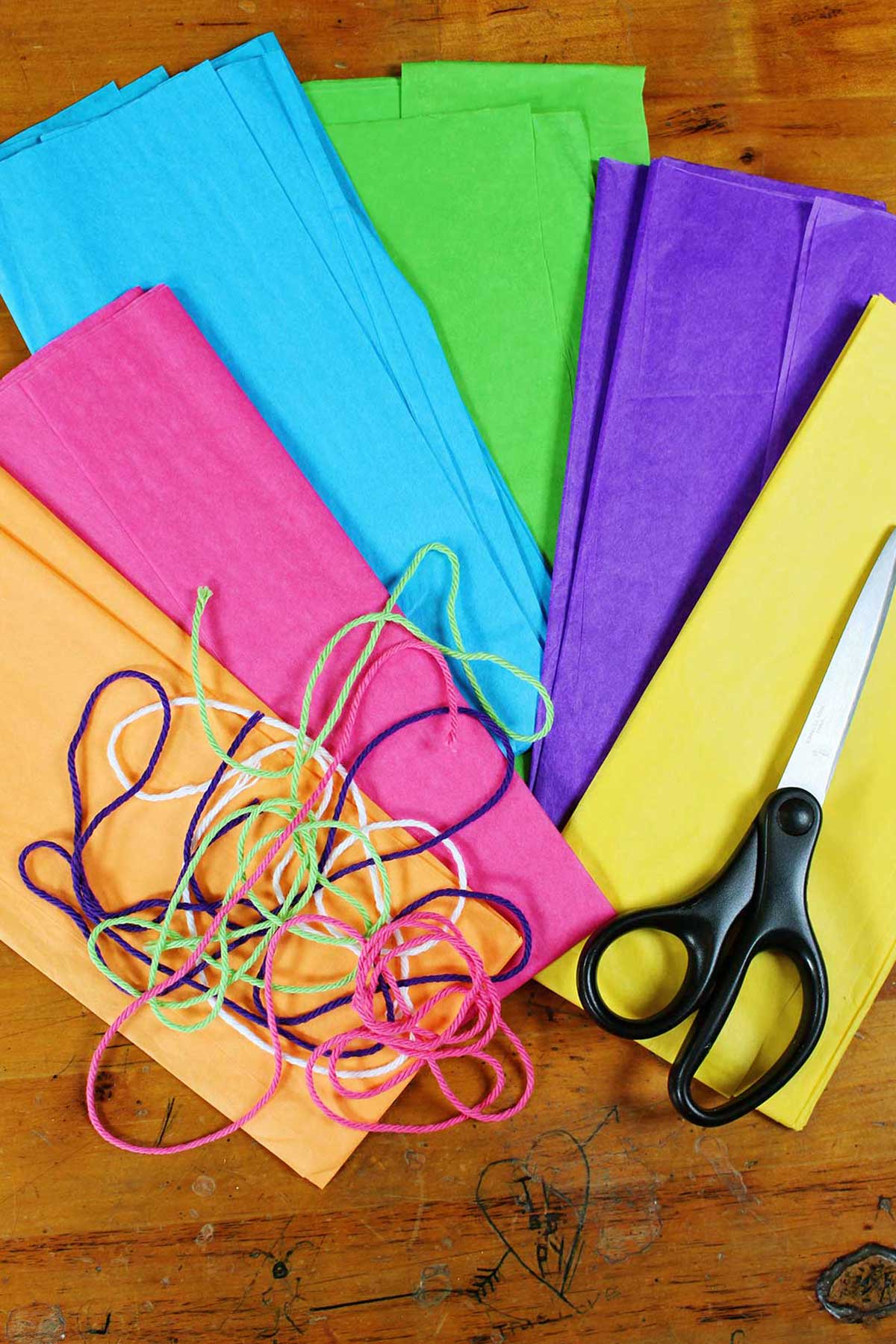Pilhas de papel de seda laranja, rosa, azul, verde, roxo e amarelo com linha e tesoura descansando sobre eles.