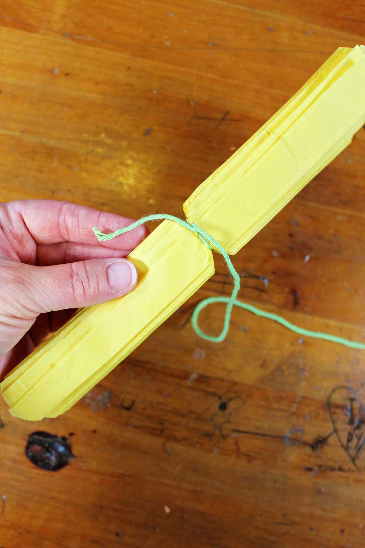 Mão segurando papel crepom amarelo dobrado e aparado com um fio verde amarrado ao redor do centro.