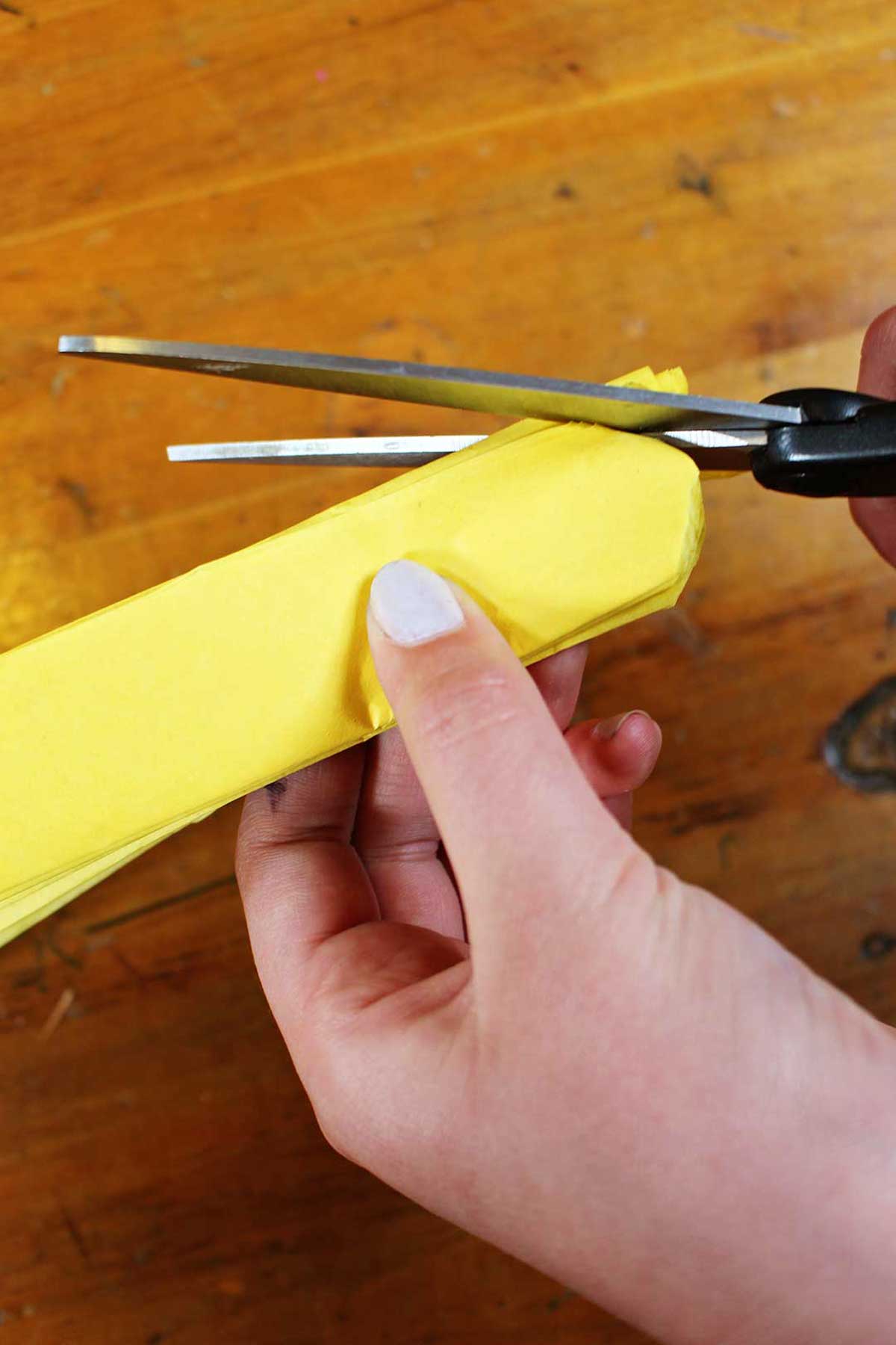 Uma mão cortando as extremidades do papel crepom amarelo dobrado para uma borda arredondada com tesoura.