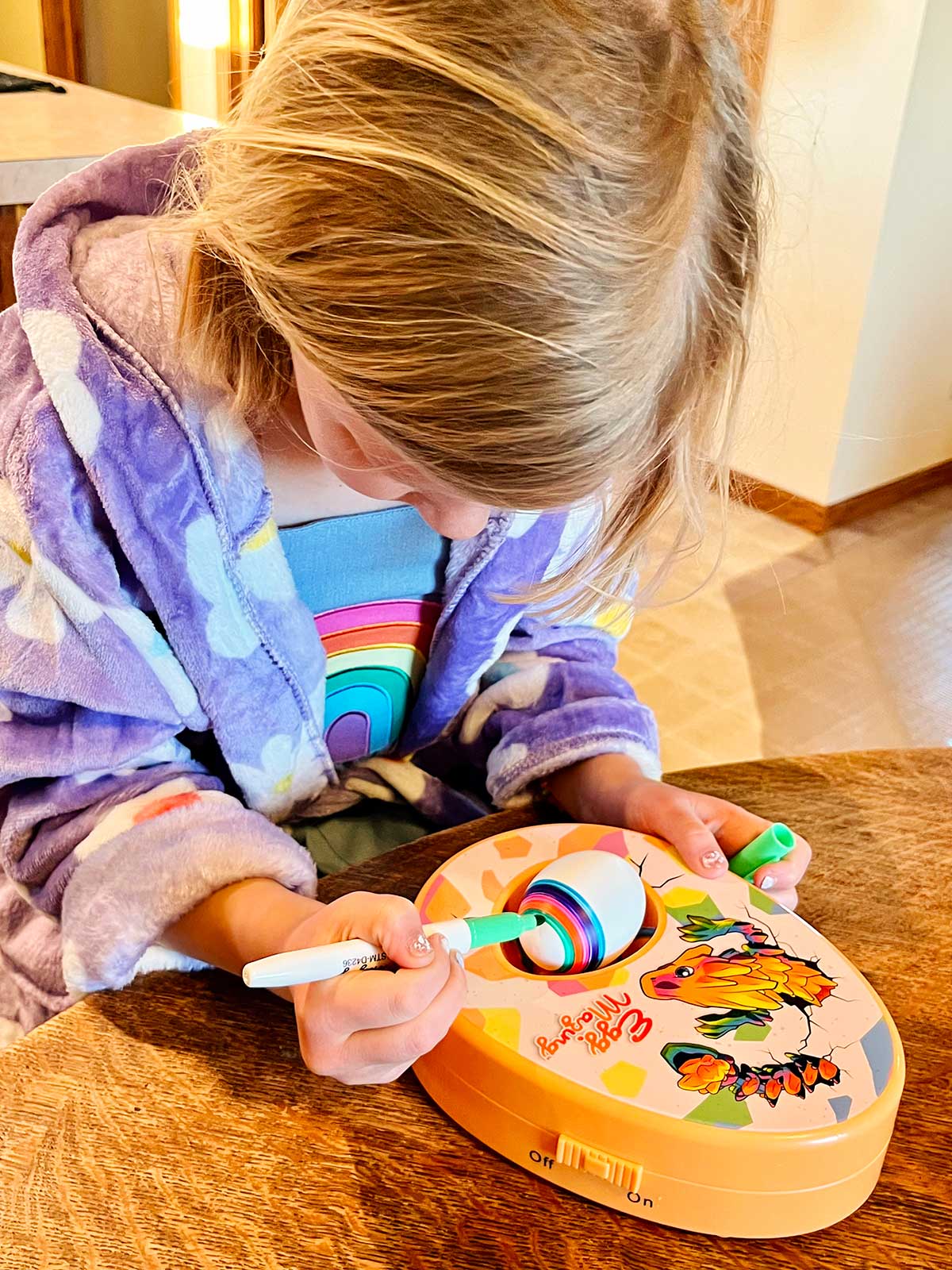 Uma menina jovem com cabelo loiro e um roupão roxo florido colorindo um ovo em uma mesa de cozinha.