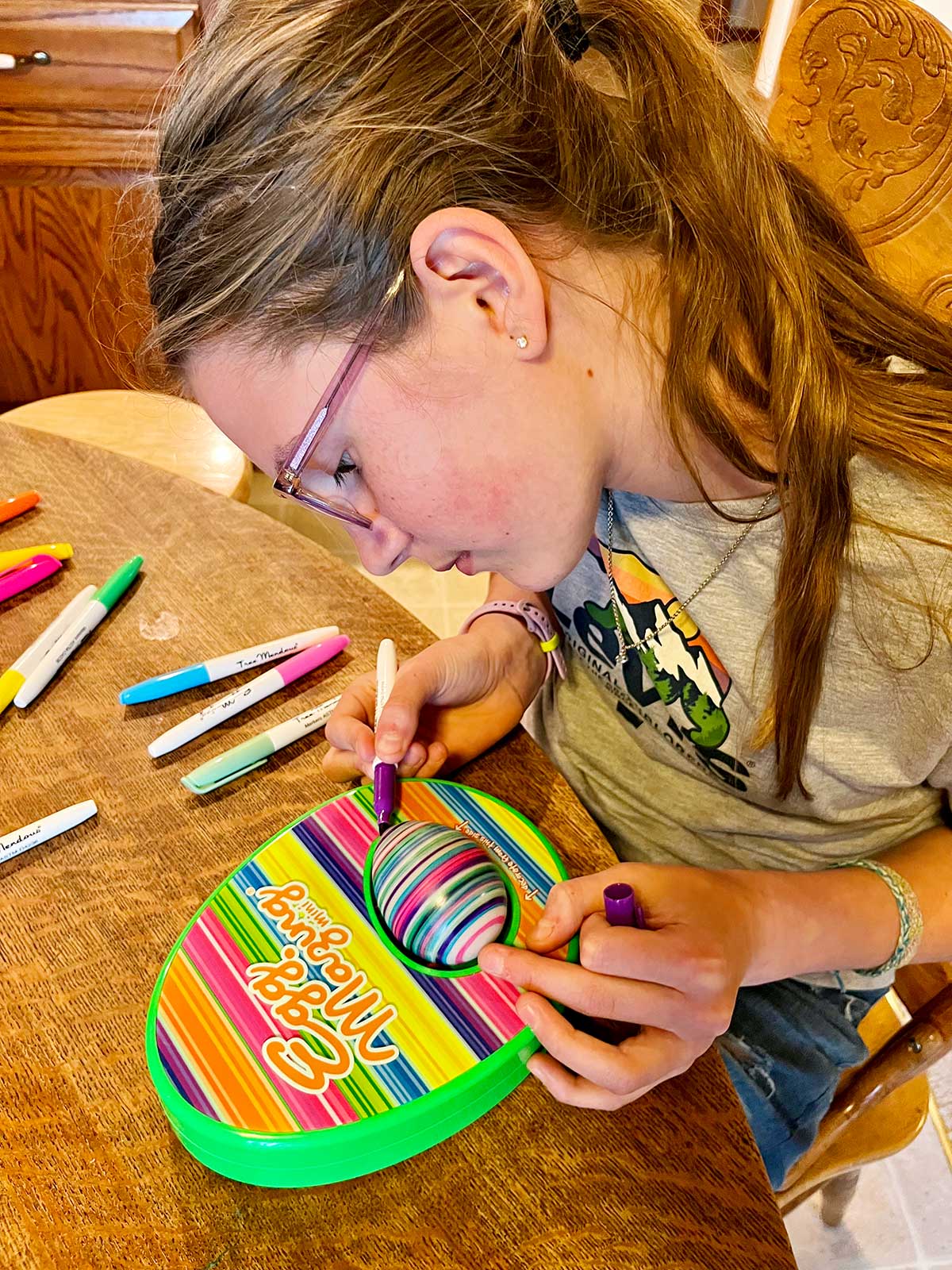 Uma menina em idade escolar com cabelo castanho claro colorindo um ovo com uma máquina EggMazing em uma mesa de cozinha.