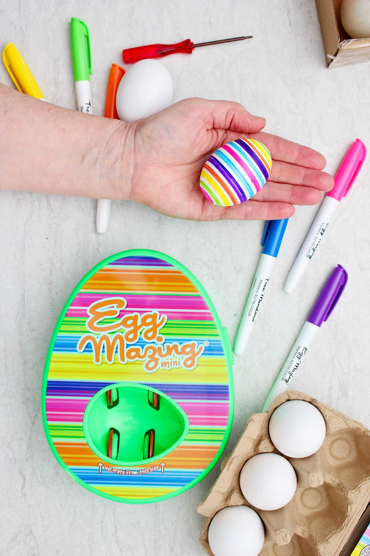 Uma pessoa segurando um ovo colorido listrado ao lado do kit EggMazing listrado.
