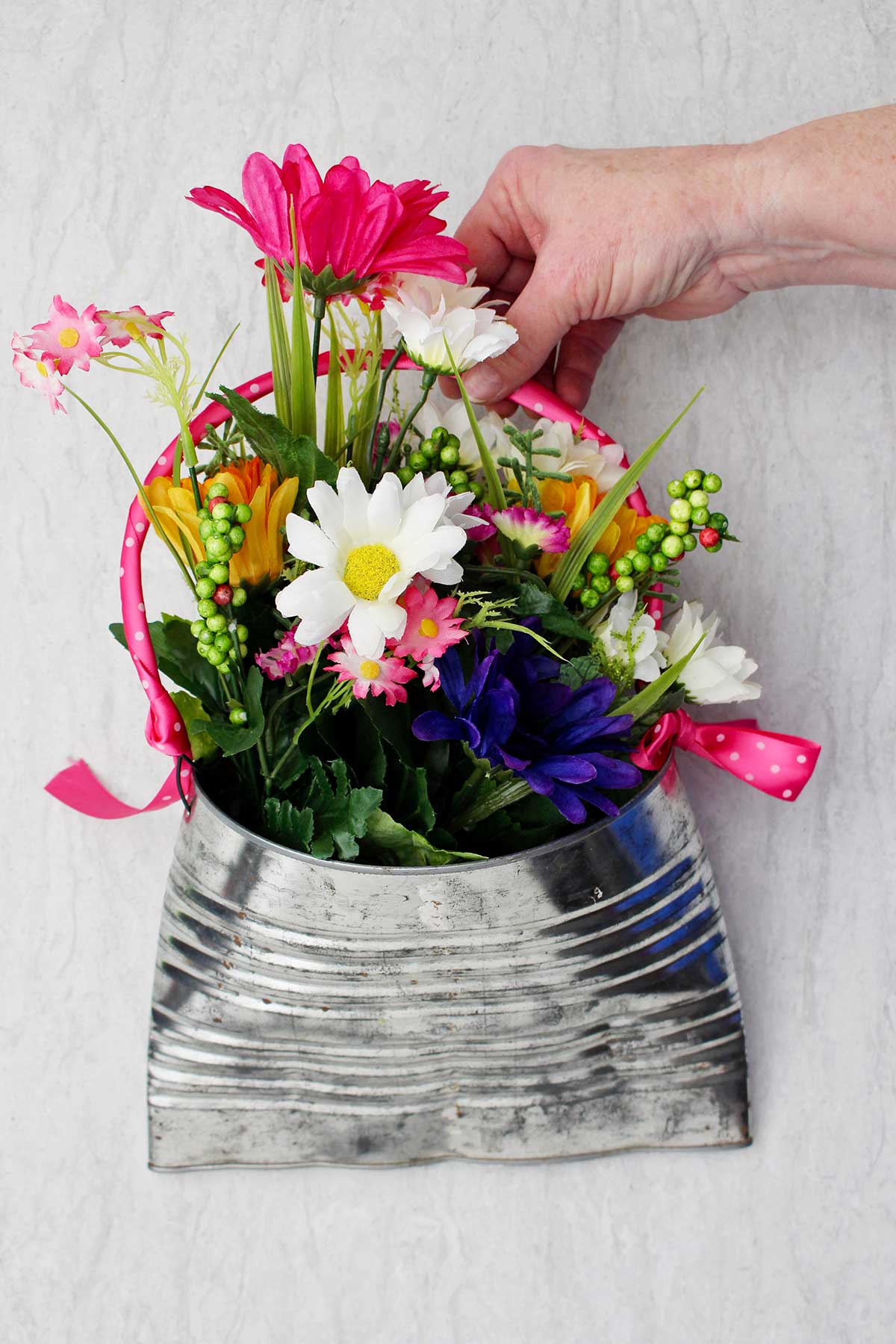 Uma pessoa segurando um vaso de lata reciclado completo, cheio de flores rosas, brancas e roxas e folhagens.