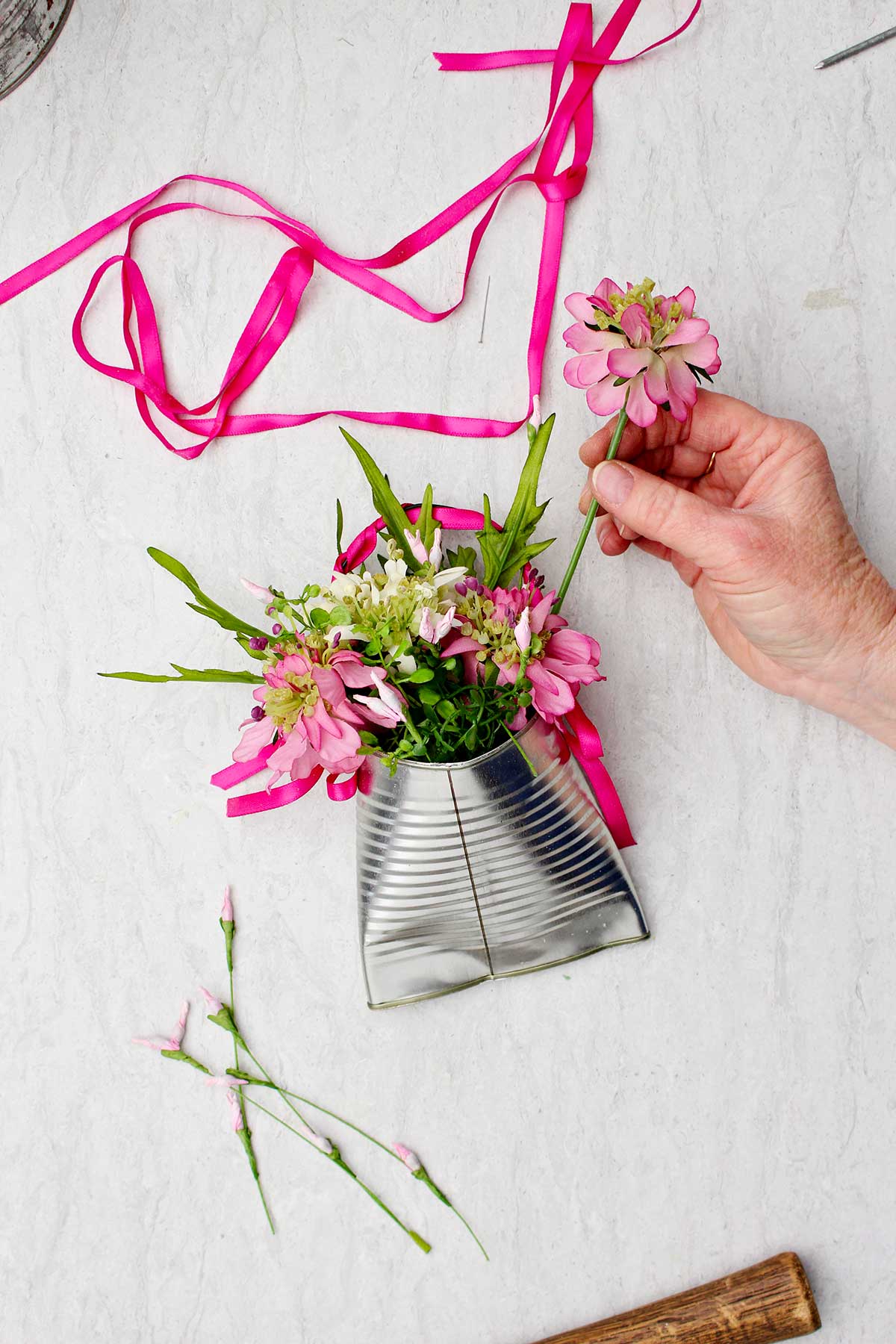 Uma pessoa colocando uma flor rosa em um vaso de lata reciclado completo com fita rosa extra ao lado.