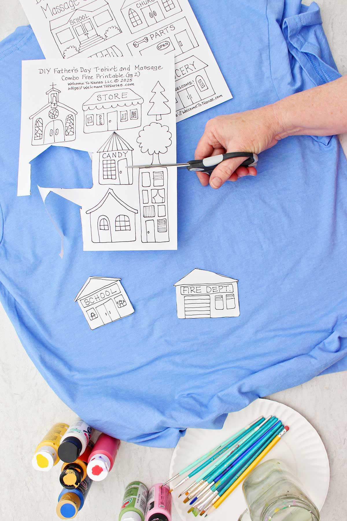 Uma pessoa cortando moldes de diferentes edifícios para a camiseta do Dia dos Pais sobre uma camiseta azul com outros suprimentos próximos.