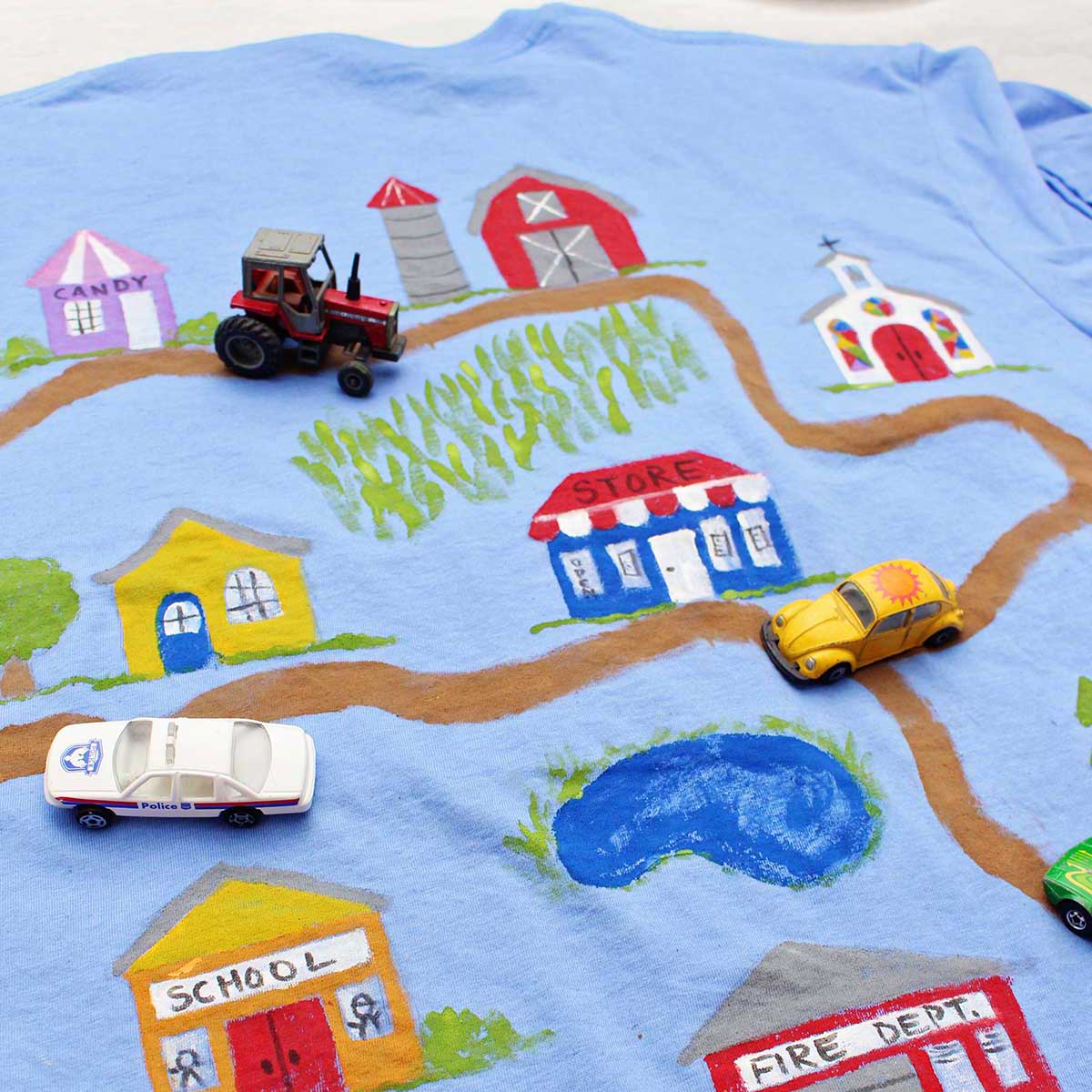 Um carro de polícia, trator e carrinhos de brinquedo Volkswagen Beetle amarelo em uma camiseta do Dia dos Pais pintada com uma estrada de terra levando a vários edifícios.