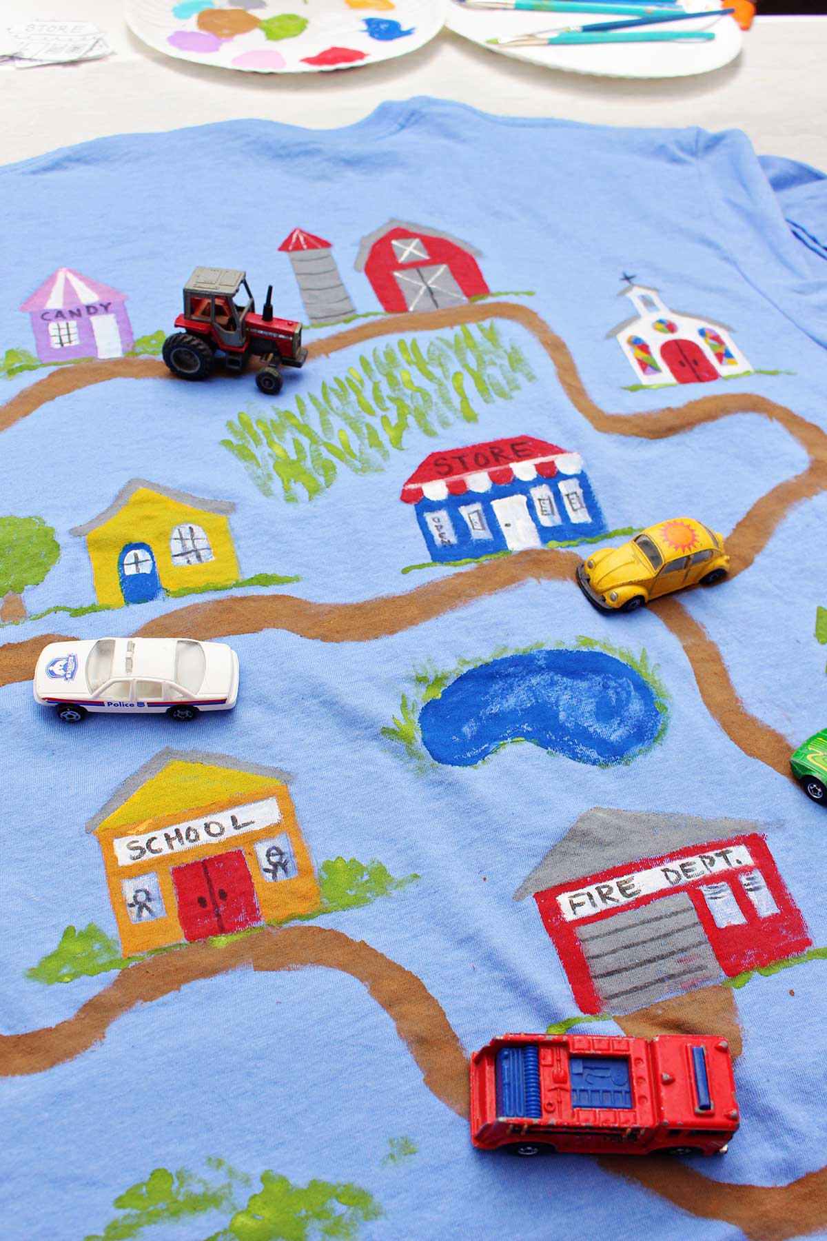 Um carro de polícia, caminhão de bombeiros, trator e carrinhos de brinquedo Volkswagen Beetle amarelo em uma camiseta do Dia dos Pais pintada com uma estrada de terra levando a vários edifícios.