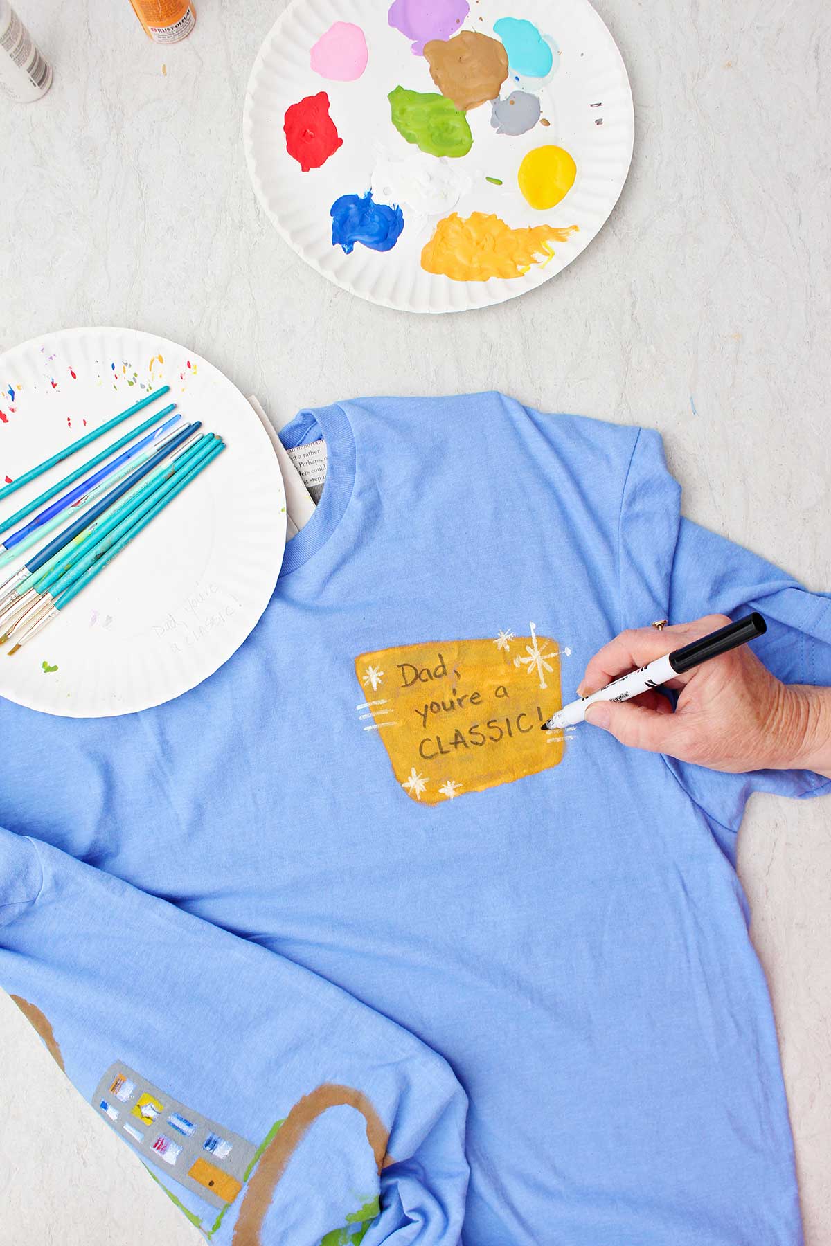 Uma pessoa escrevendo 'Pai, você é um clássico' na frente de uma camiseta azul com um prato de tinta e pincéis por perto.