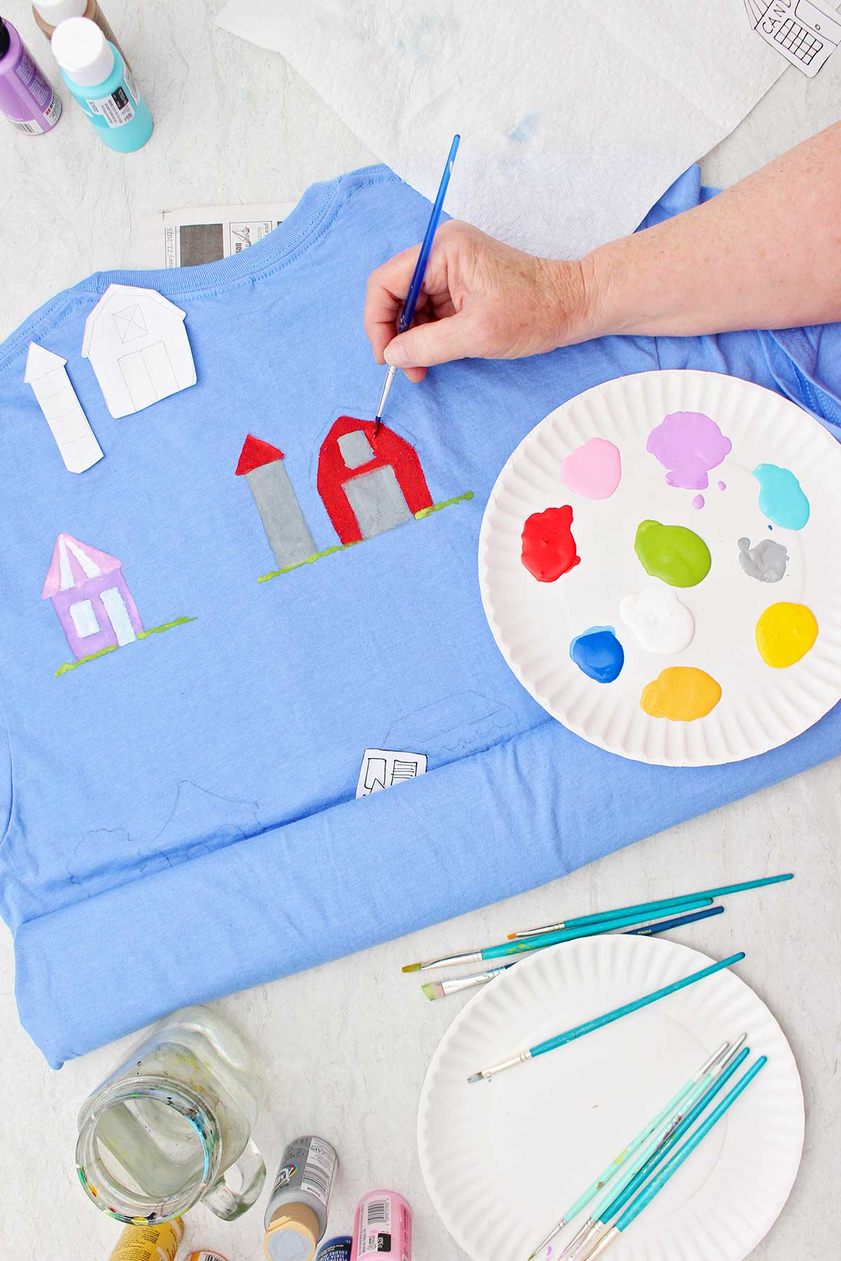 Uma pessoa pintando um celeiro de vermelho em uma camiseta azul com um prato de tinta e outros suprimentos por perto.