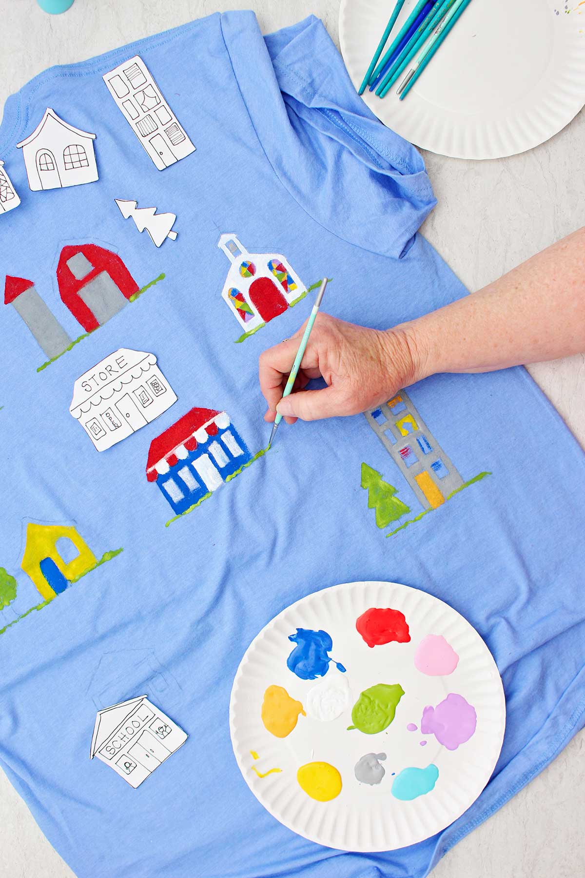 Uma pessoa pintando detalhes de grama perto de uma loja pintada em uma camiseta azul com um prato de tinta por perto.