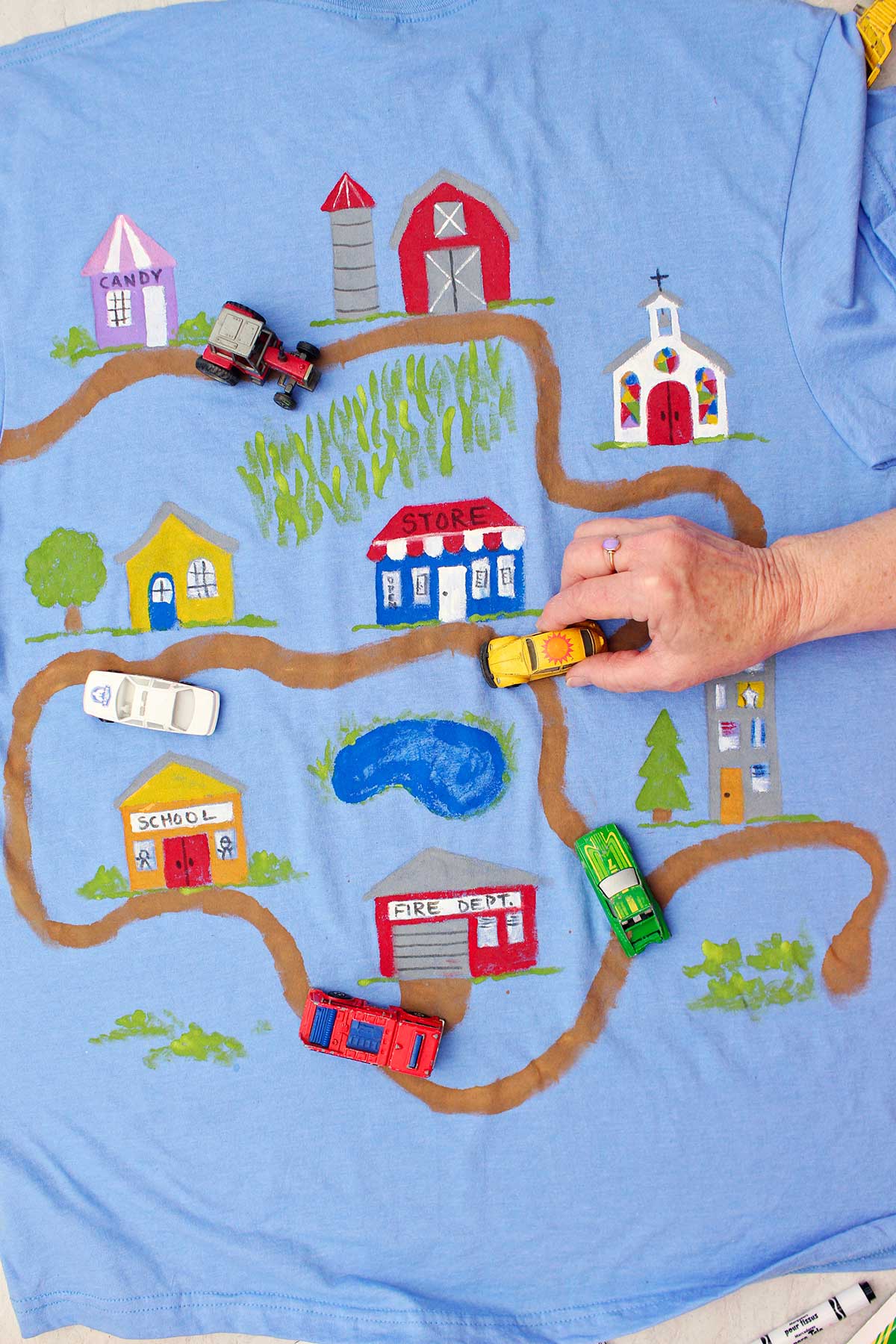 Pessoa dirigindo um carro de brinquedo Volkswagen Beetle amarelo em uma camiseta do Dia dos Pais pintada com uma estrada de terra levando a vários edifícios da cidade.