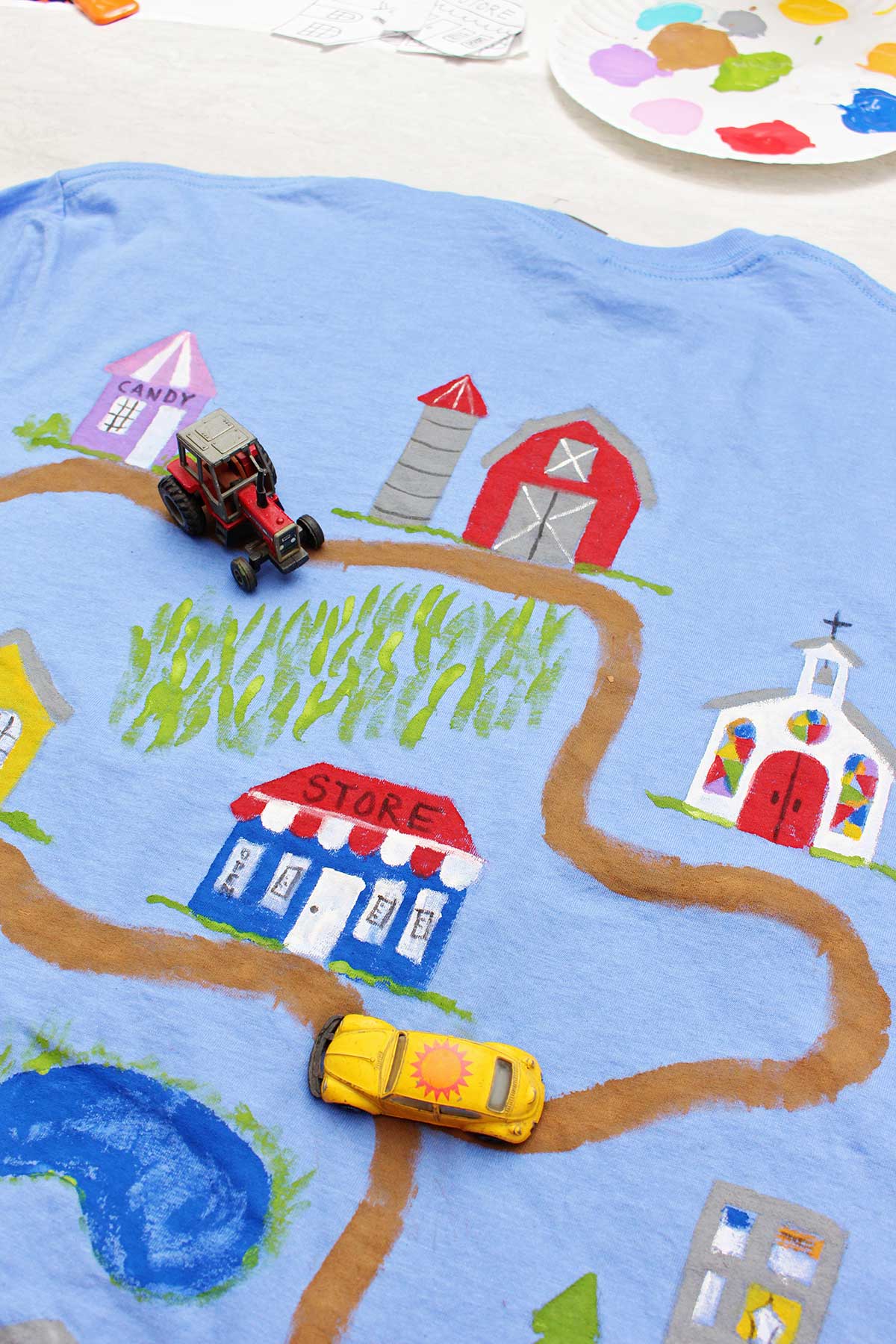 Um trator e um carro de brinquedo Volkswagen Beetle amarelo em uma camiseta do Dia dos Pais pintada com uma estrada de terra levando a vários edifícios com um prato de tinta ao fundo.