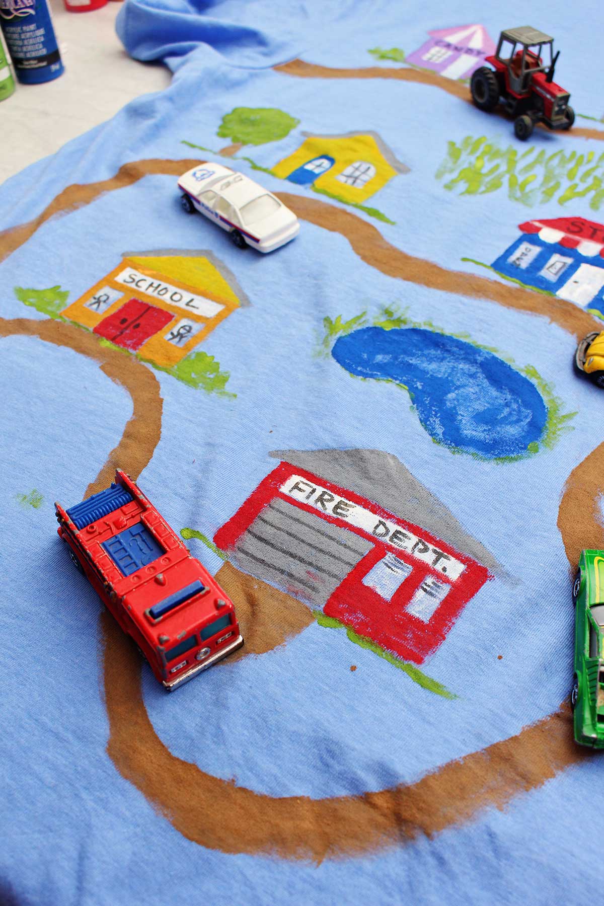Um carro de polícia, trator e caminhão de bombeiros de brinquedo em uma camiseta do Dia dos Pais pintada com uma estrada de terra levando a vários edifícios.