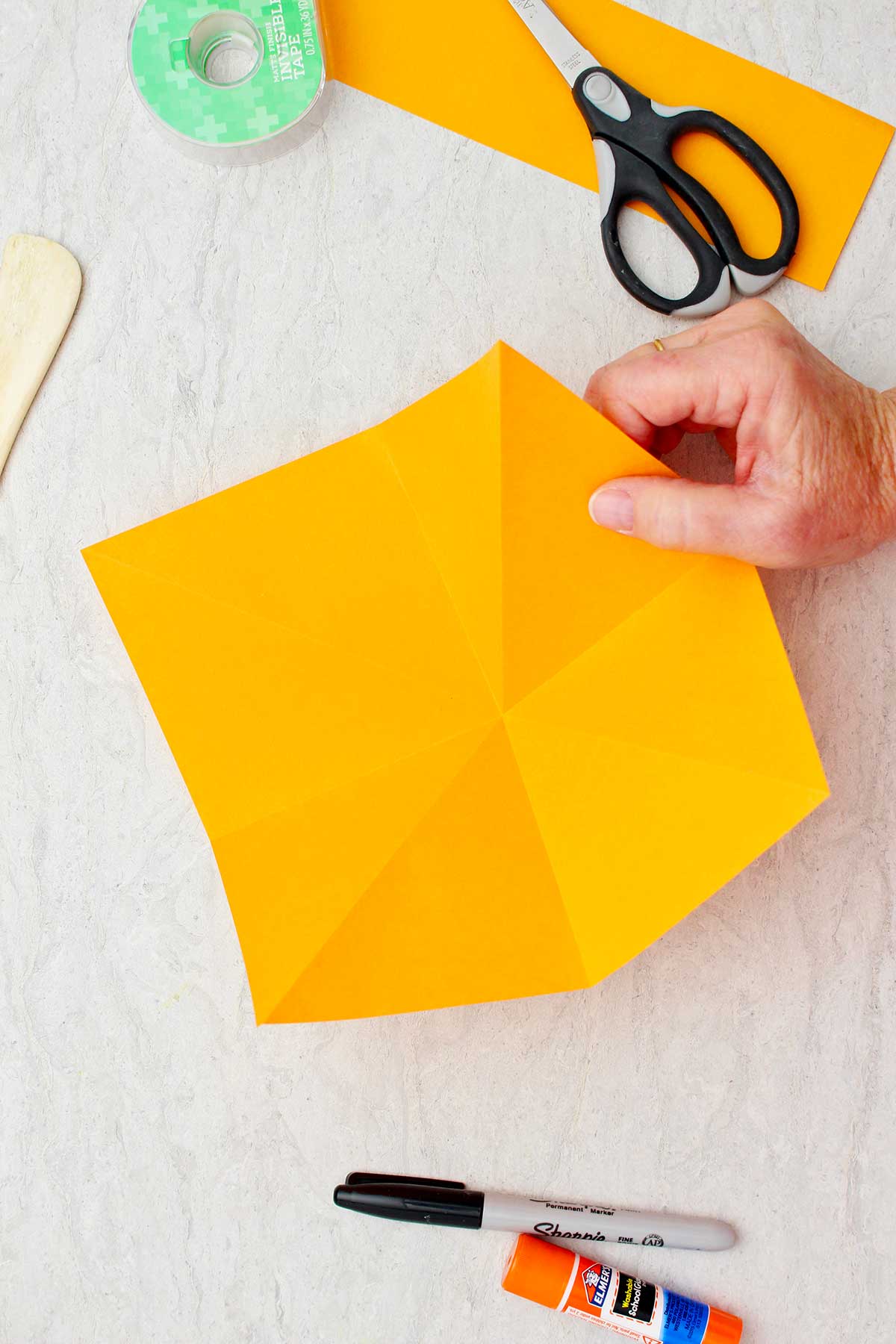 Mão segurando o lado direito de um pedaço cortado de papel laranja pronto para ser dobrado em uma abóbora de origami.