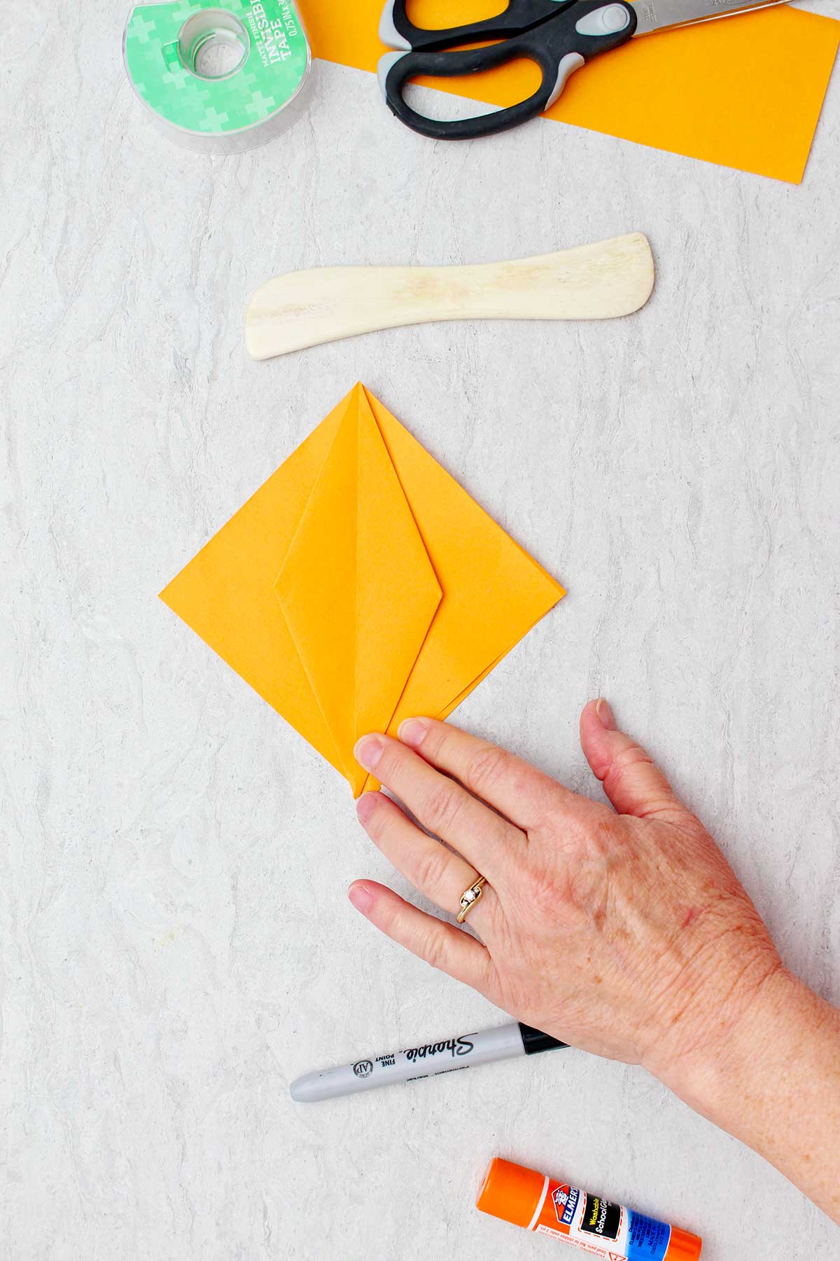 Um pedaço quadrado de papel laranja com um diamante dobrado no centro e outros materiais por perto para criar abóboras de origami.