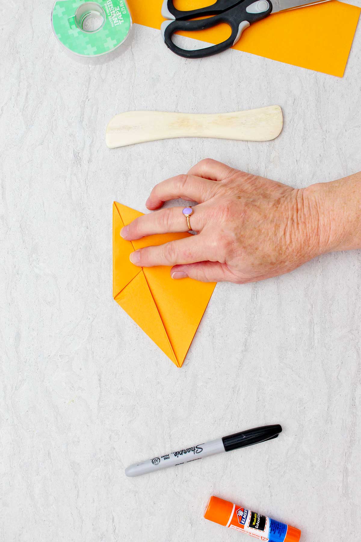 Pessoa dobrando um pedaço de papel laranja para criar uma abóbora de origami com outros materiais por perto.