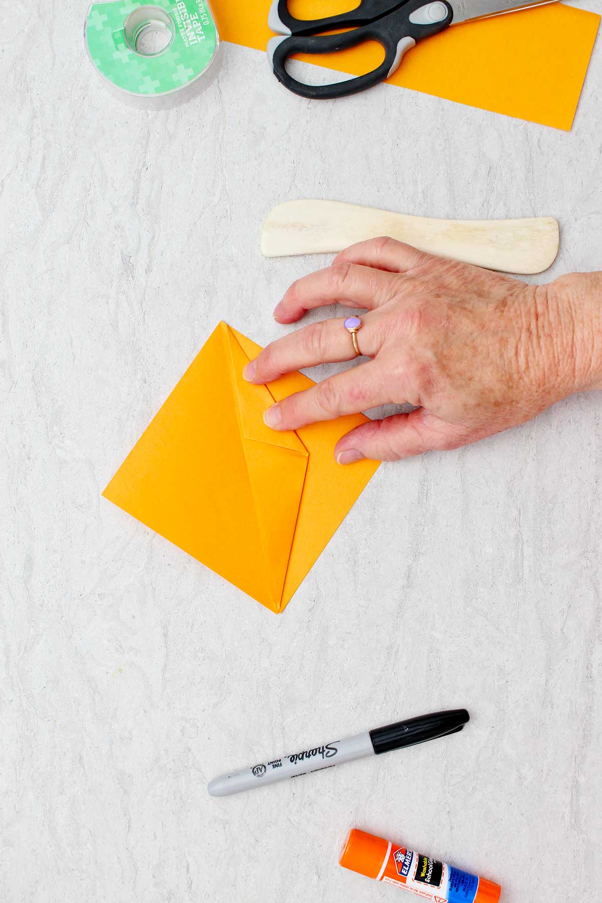 Pessoa dobrando um pedaço de papel laranja para criar uma abóbora de origami com outros materiais por perto.