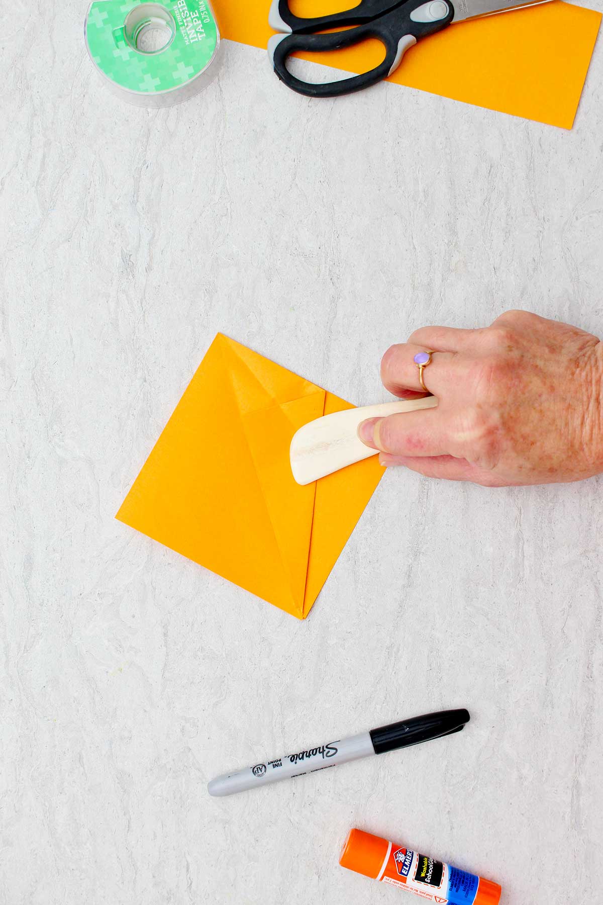 Pessoa usando um bastão de madeira arredondado para afiar as bordas dobradas de um pedaço de papel laranja usado para fazer uma abóbora de origami.
