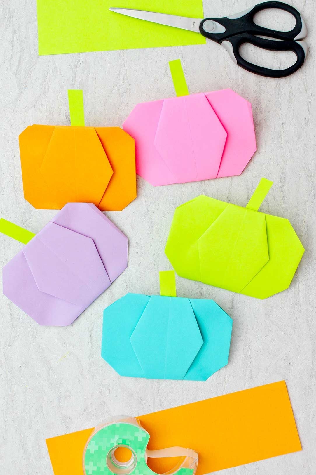 Cinco abóboras de origami de diferentes cores com tesouras, fita e outros pedaços de papel colorido por perto.