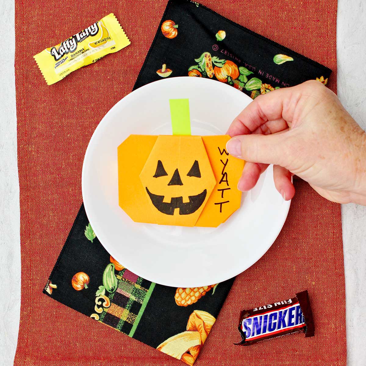 Mão colocando uma abóbora de origami laranja em um lugar para uma configuração com alguns doces de Halloween por perto.