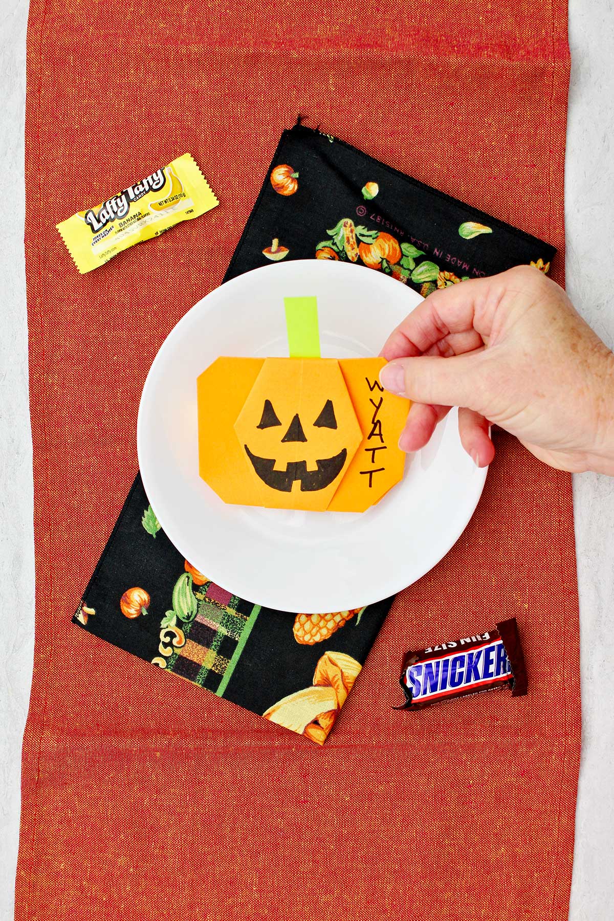 Mão colocando uma abóbora de origami laranja em um lugar para uma configuração de mesa com alguns doces de Halloween por perto.