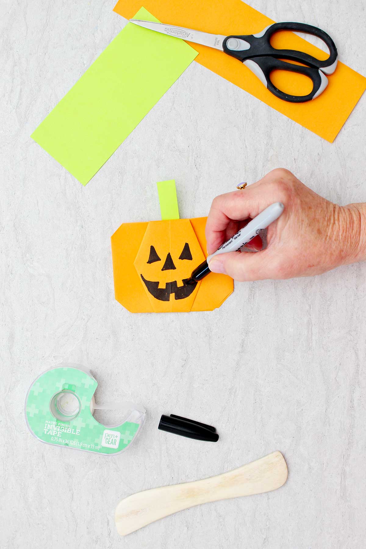 Uma pessoa desenhando um rosto de jack-o-lantern em uma abóbora de origami laranja com outros suprimentos por perto.