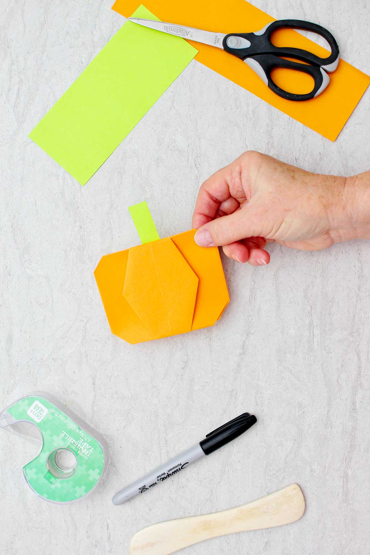 Pessoa segurando uma abóbora de origami laranja completa com um caule verde-lima com outros suprimentos para fazer abóbora de origami por perto.