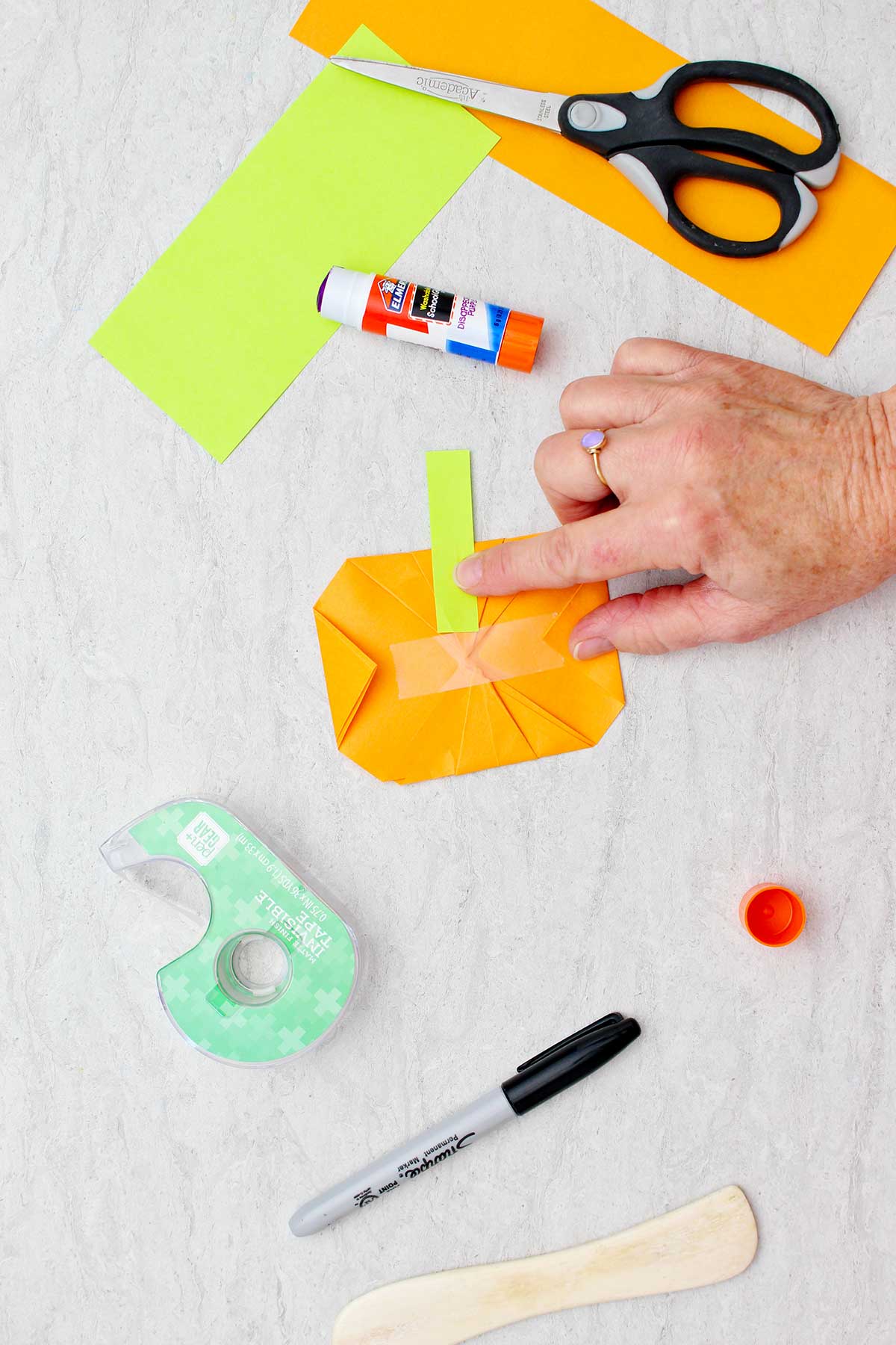 Uma pessoa colando um retângulo verde na parte de trás de uma abóbora de origami laranja como um caule, com outros suprimentos para fazer abóboras de origami por perto.
