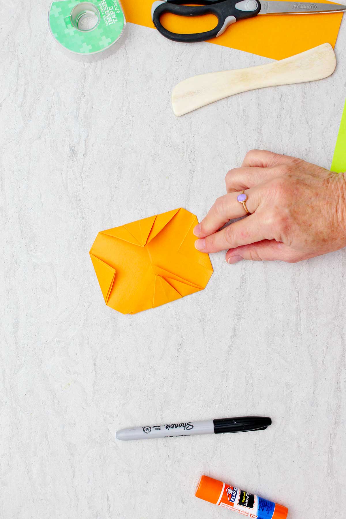 Pessoa dobrando um canto de papel laranja e mostrando o lado de trás de uma abóbora de origami quase completada.