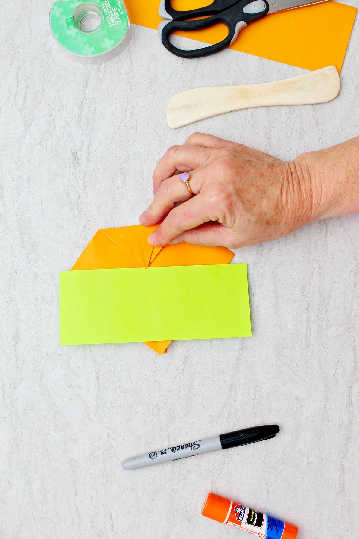 Um retângulo verde de papel descansando sobre um pedaço de papel laranja que uma pessoa está dobrando com outros suprimentos próximos para criar abóboras de origami.