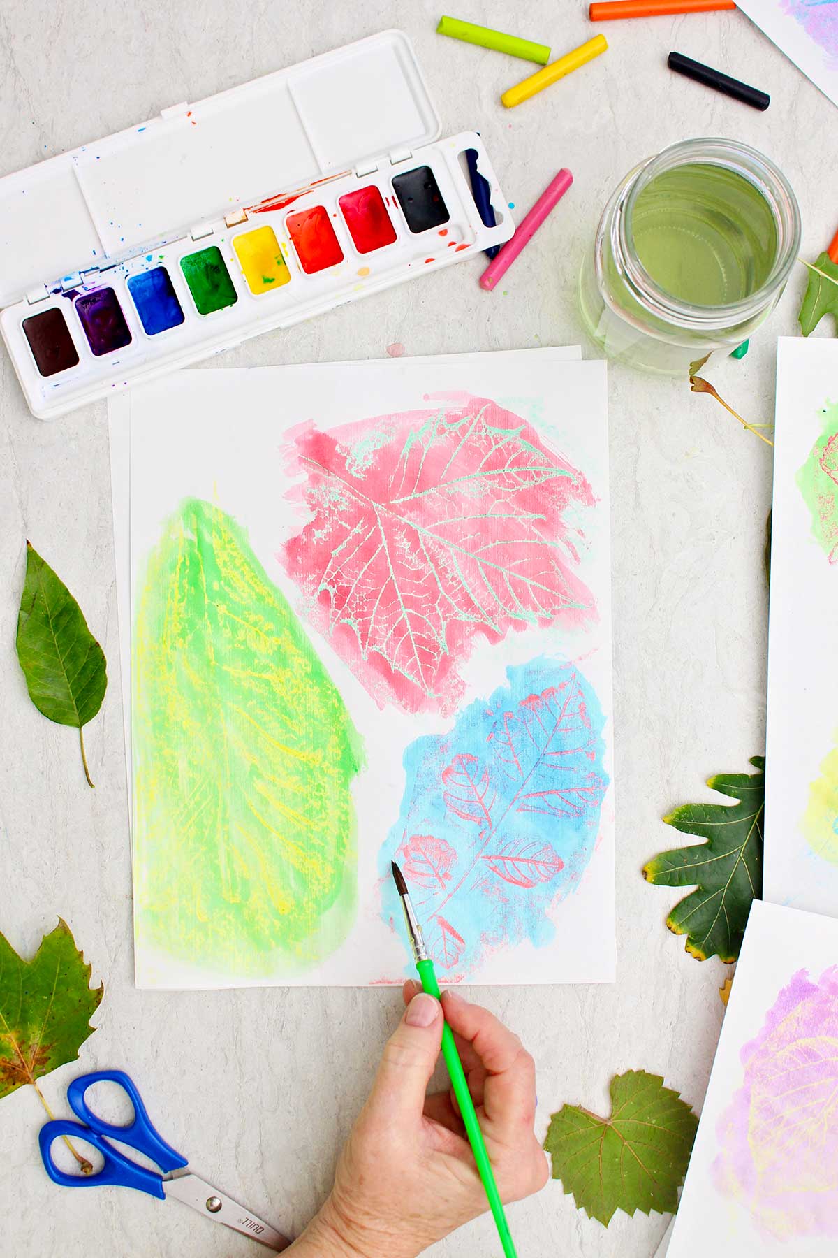 Uma pessoa pintando com aquarela sobre impressões de folhas com folhas e aquarelas espalhadas ao redor.