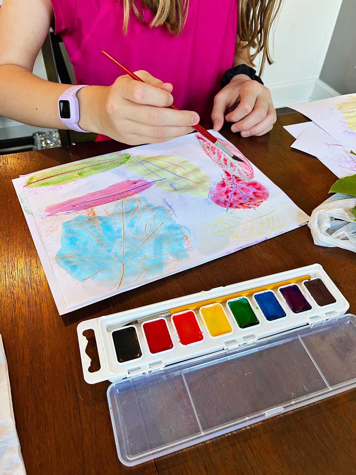 Uma menina jovem com uma camisa rosa pintando com aquarela sobre impressões de folhas em uma mesa.