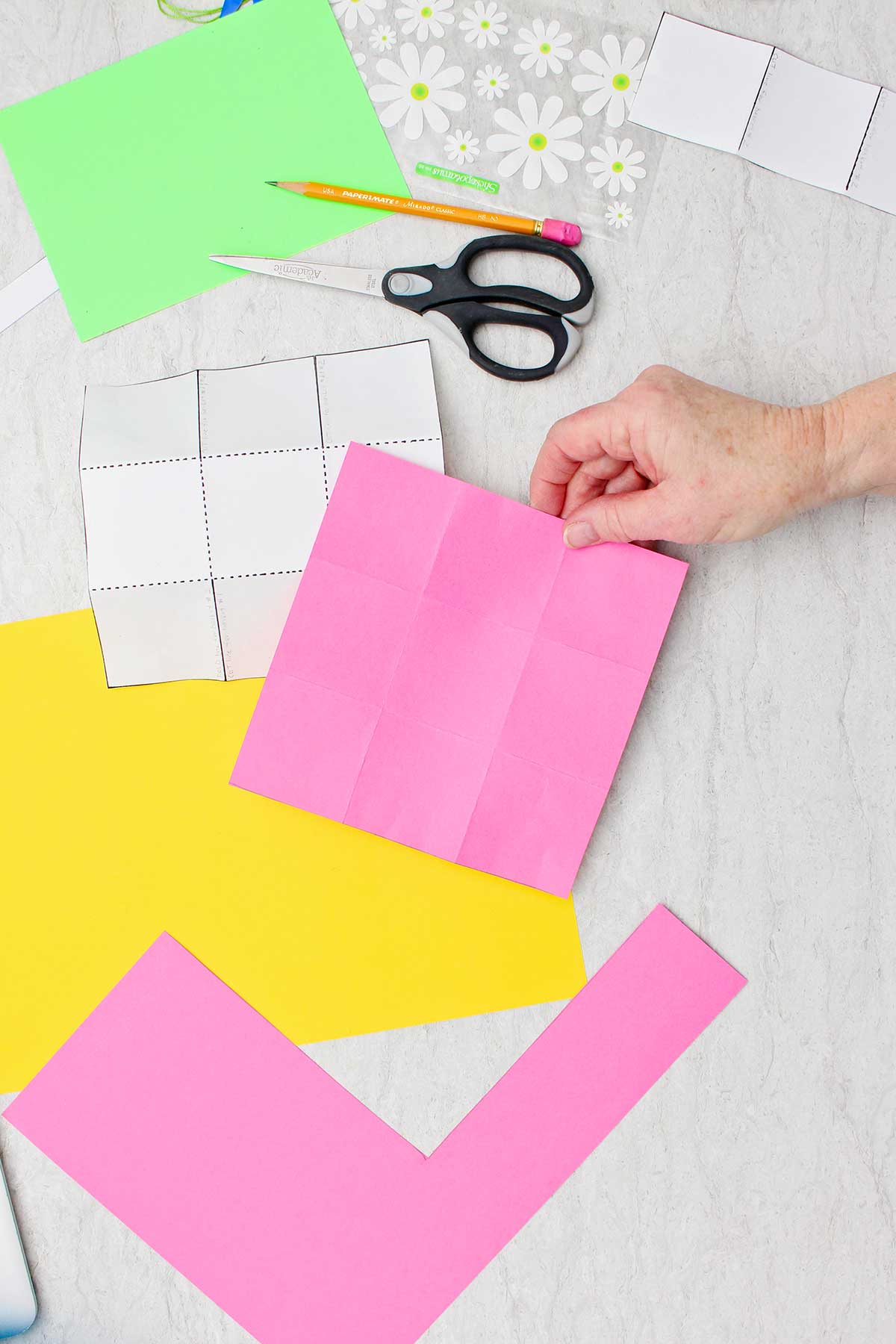Uma pessoa segurando um pedaço quadrado de papel rosa com linhas de dobra para criar nove caixas iguais com outros suprimentos ao redor.