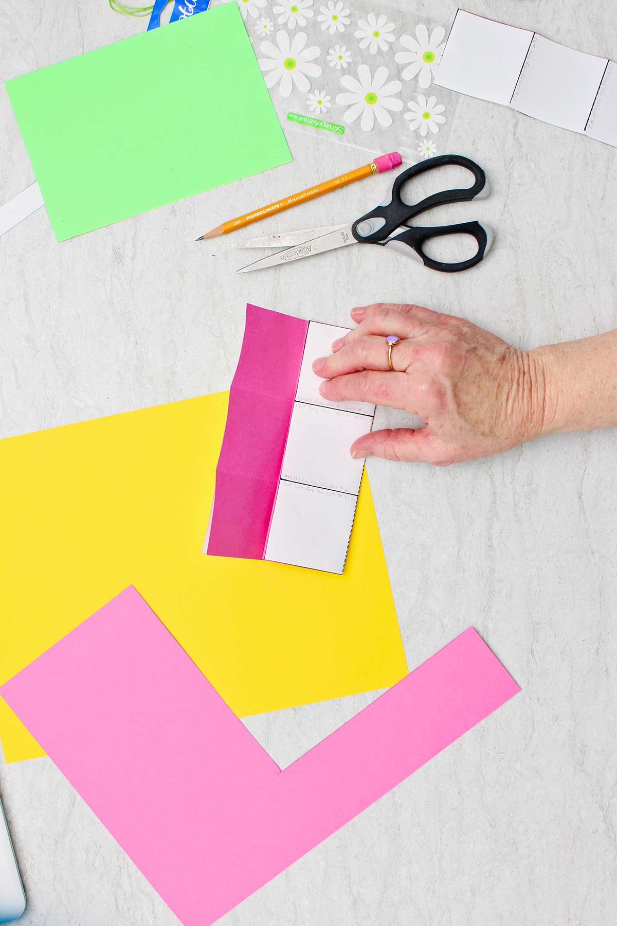 Pessoa segurando um molde recortado próximo a um pedaço de papel rosa escuro para verificar linhas de dobra com outros materiais próximos.