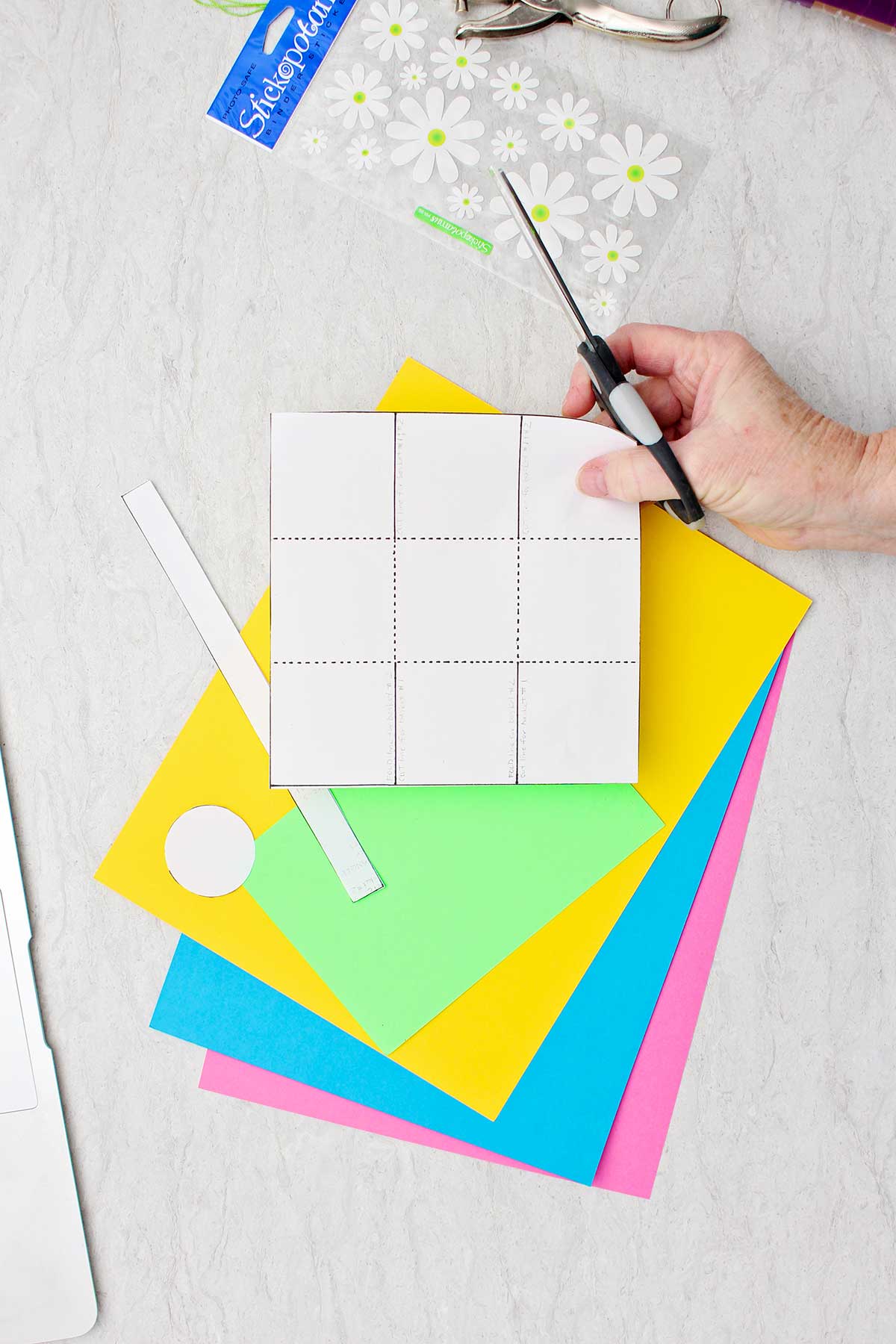 Pessoa segurando um molde para um mini cesto de Páscoa sobre uma pilha de papel colorido com uma folha de adesivos de flores próxima.