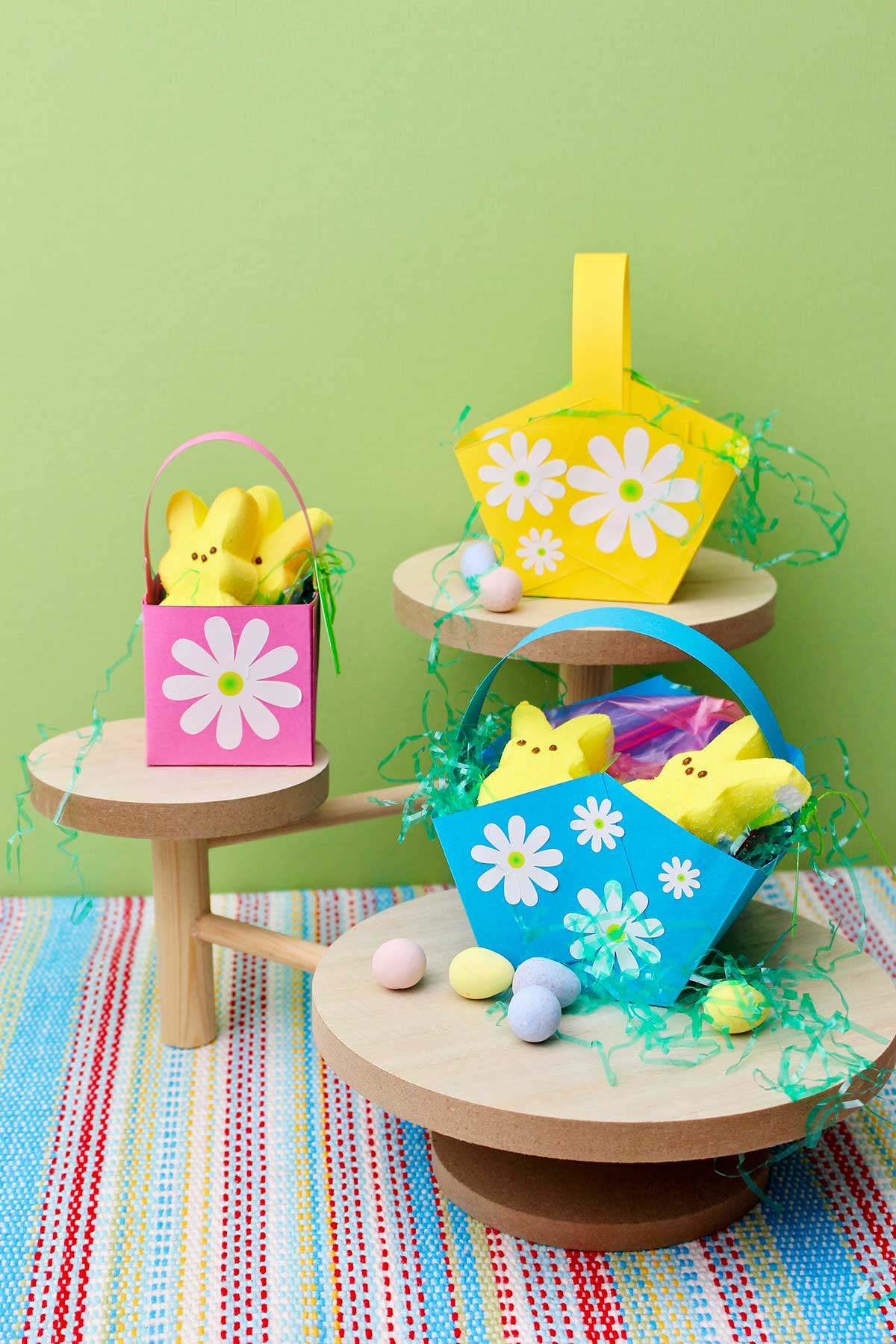 Três mini cestos de Páscoa cheios de doces com flores brancas decoradas estão sobre pedestais de madeira contra um fundo verde.