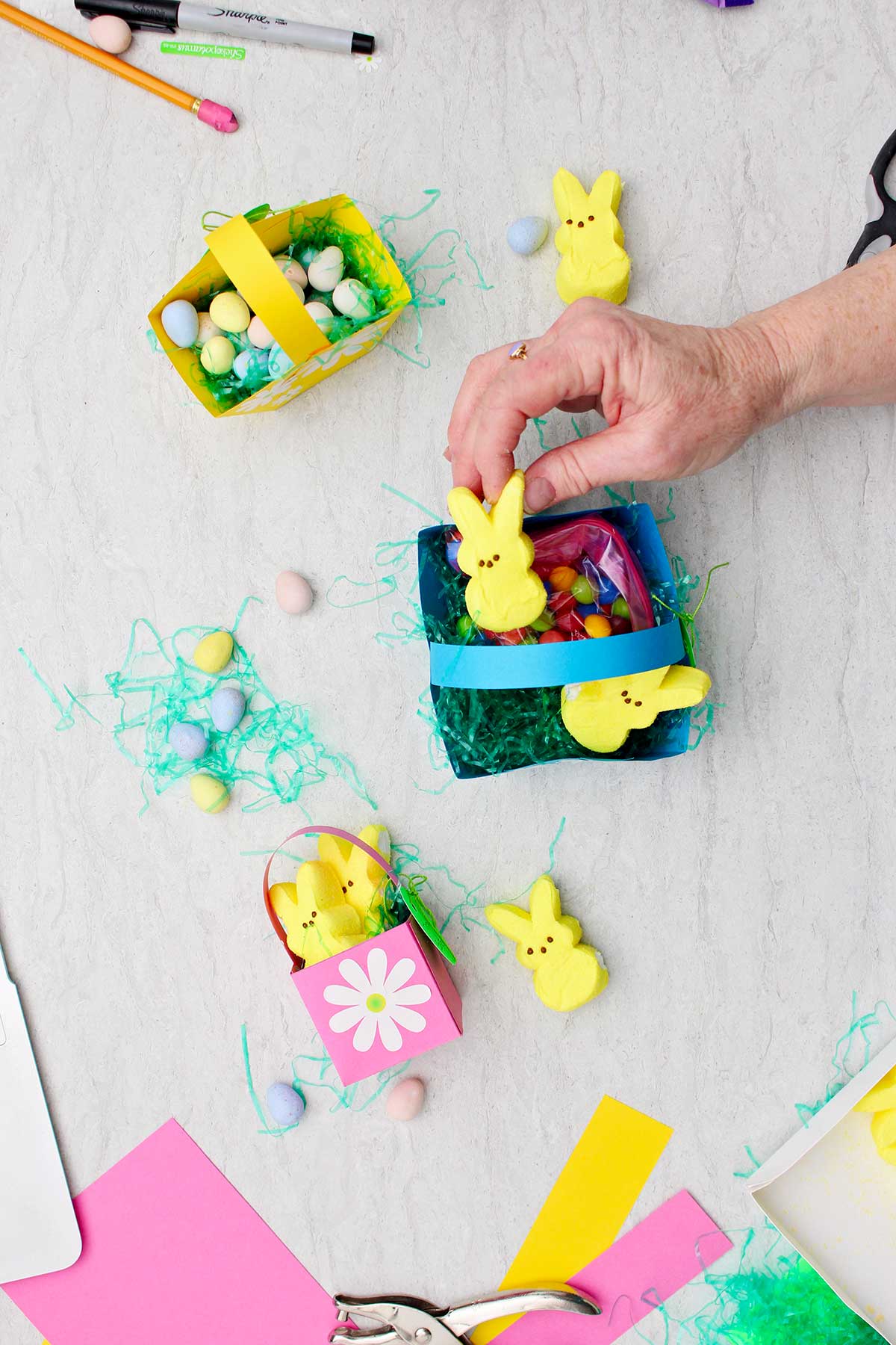 Pessoa colocando um Coelhinho de Marshmallow amarelo em uma mini cesta de Páscoa azul cheia de doces e grama de Páscoa, com outras mini cestas de Páscoa finalizadas ao lado.