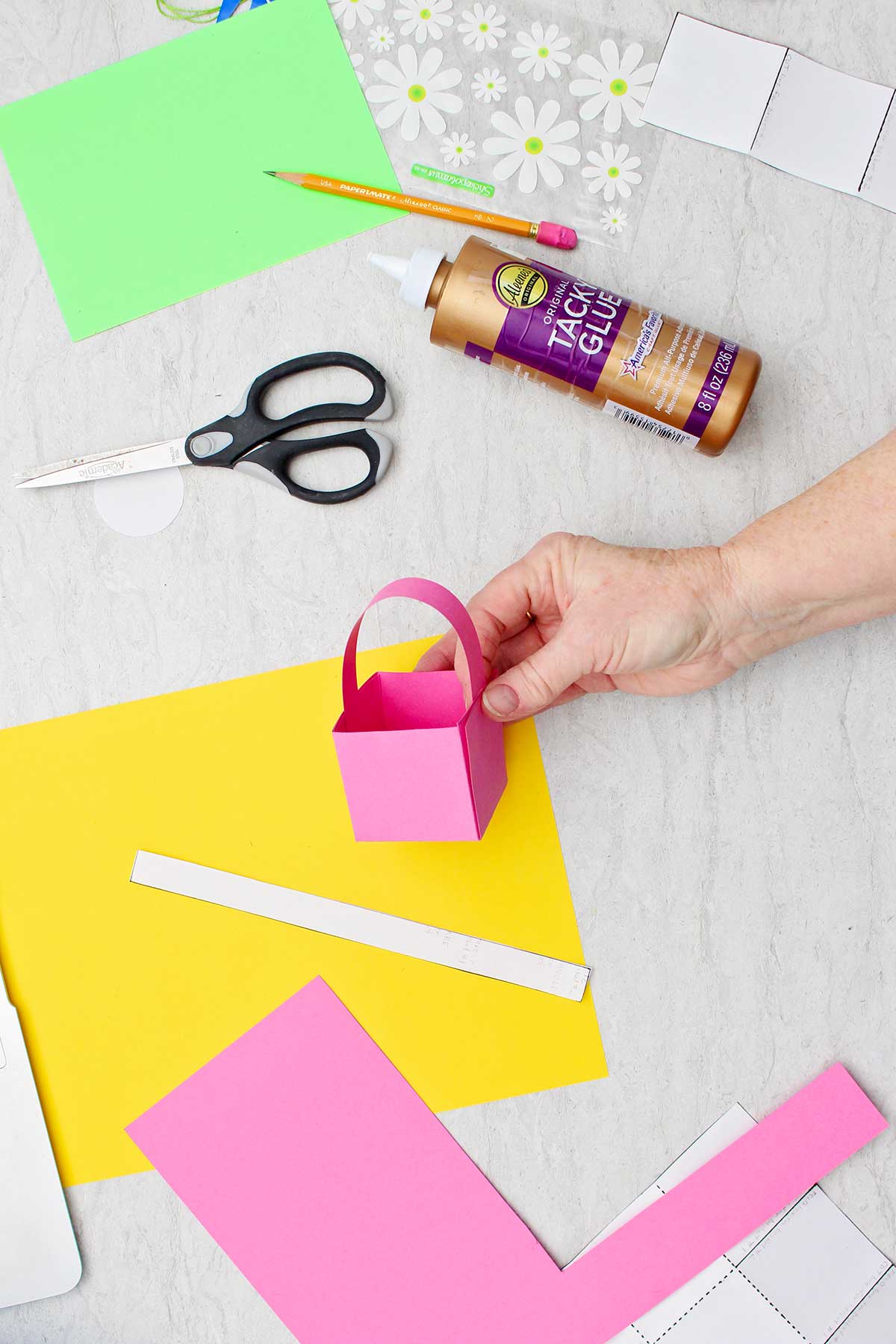 Pessoa colando a alça em um mini cesto de Páscoa rosa de papel com outros suprimentos ao redor.